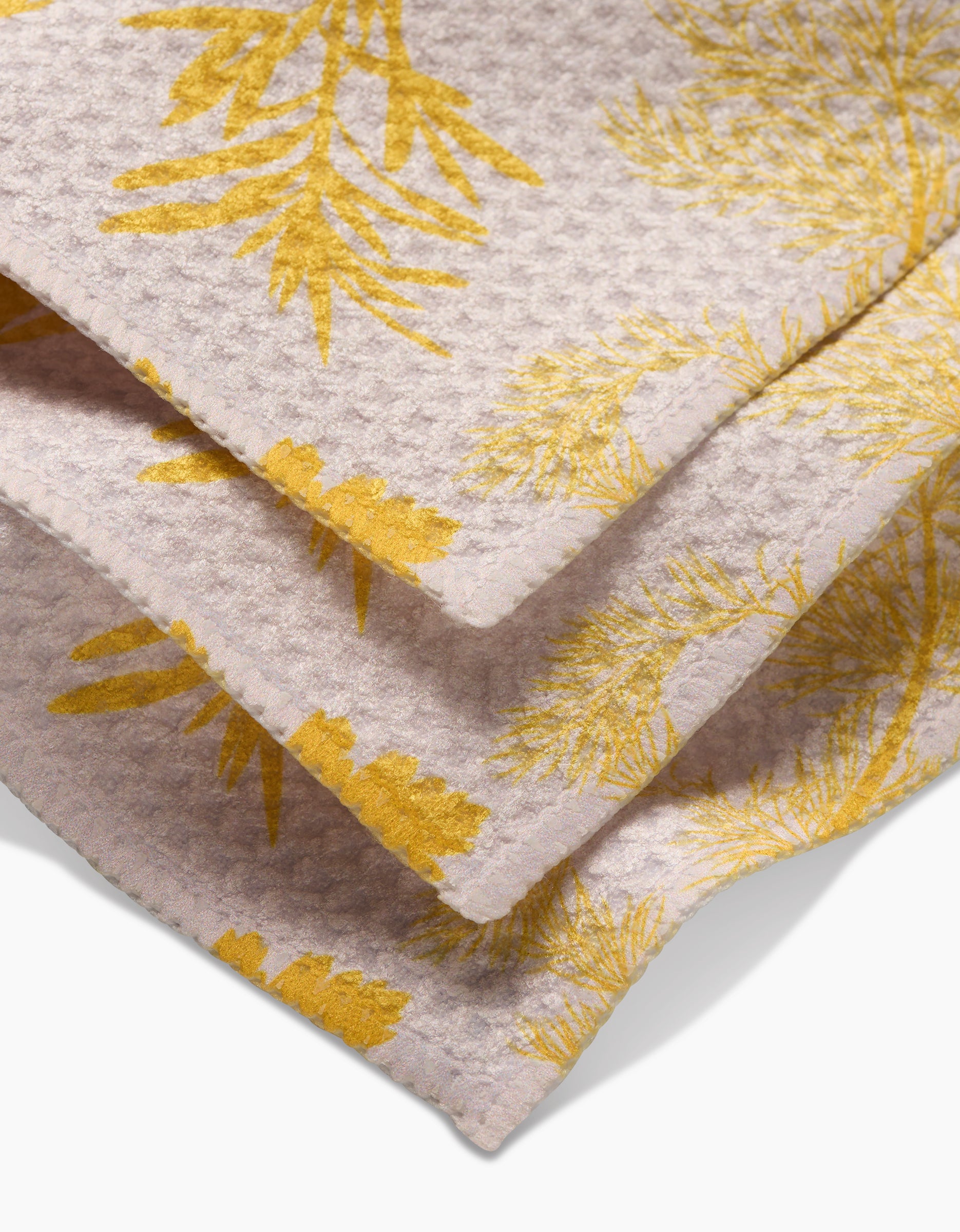 A close-up of GEOMETRYs Garden of Gold Geoweave dishcloth shows its textured waffle pattern and yellow botanical leaf prints on a pale background, all crafted from recycled materials.