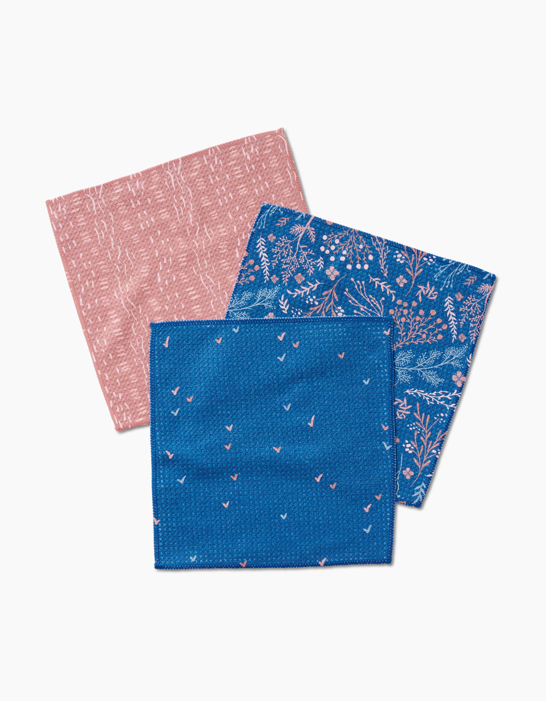 Three folded Garden Seeds napkins by Danelys Sidron: one pink with a subtle pattern, one blue with a botanical motif, and one blue featuring small pink bird-like shapes, all crafted from Geoweave fabric and displayed on a white background.