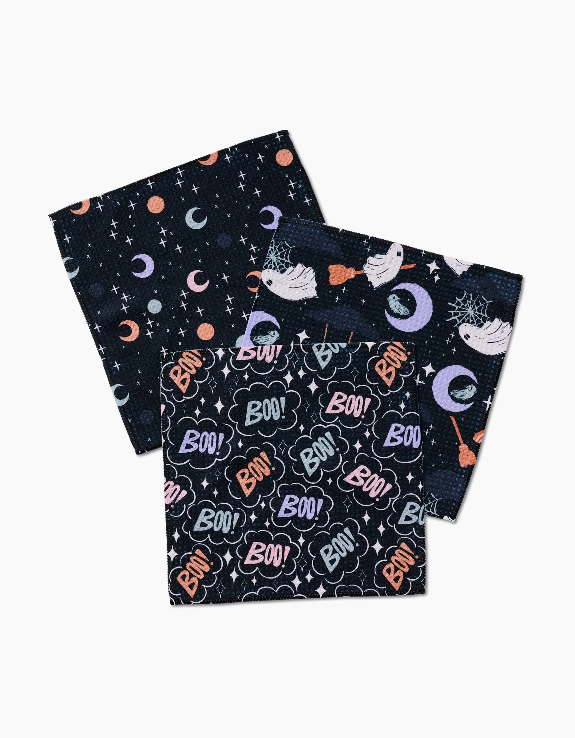 Three folded dark dishcloths from Charlie Rowan Designs’ Ghosts In The Night set—featuring moons and stars, witch hats with brooms, and colorful “Boo!” text—each crafted from recycled materials.