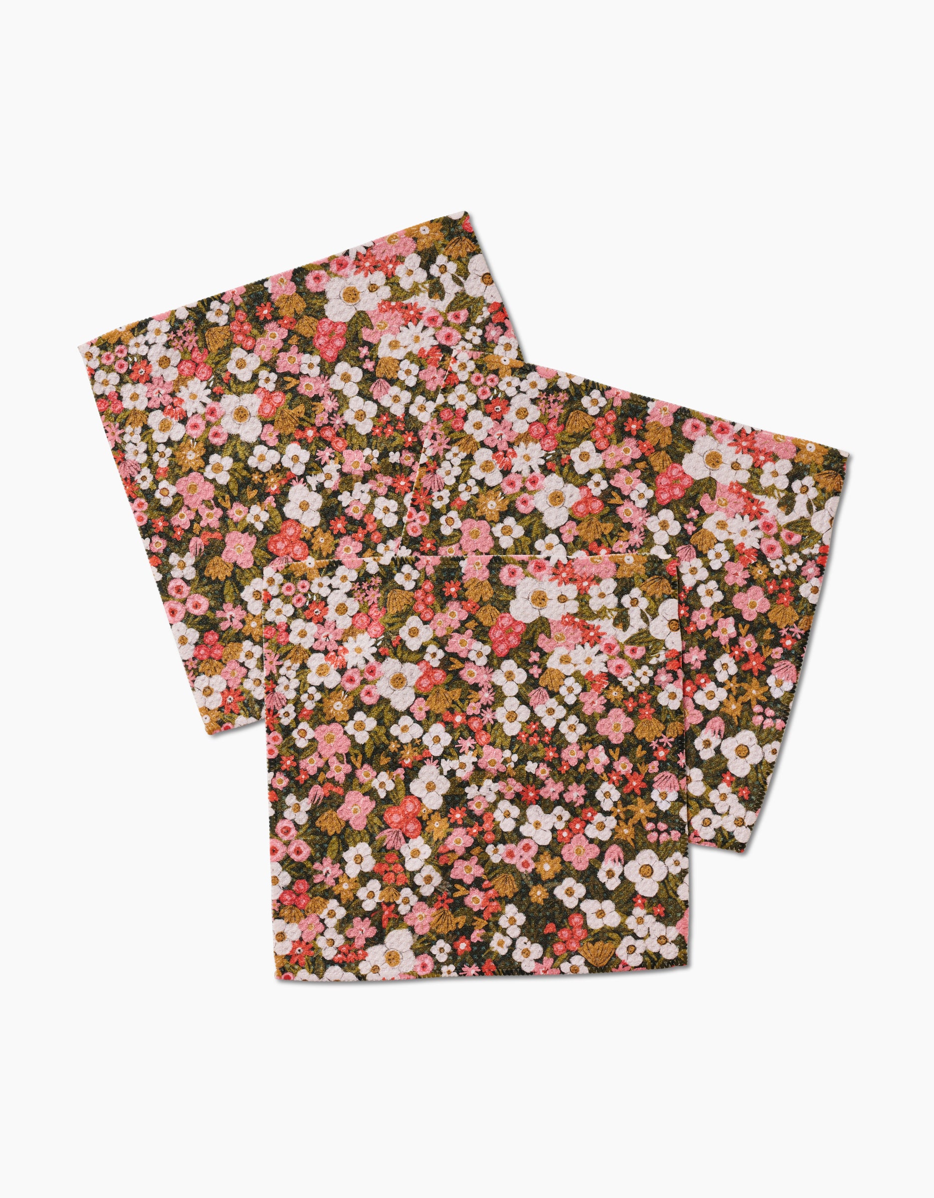 Two Gilded Garden square cloth napkins by Lisa Nelson, crafted from recycled materials, showcase a dense floral pattern with small pink, white, and orange flowers plus green leaves on a white background. The napkins are slightly overlapping.