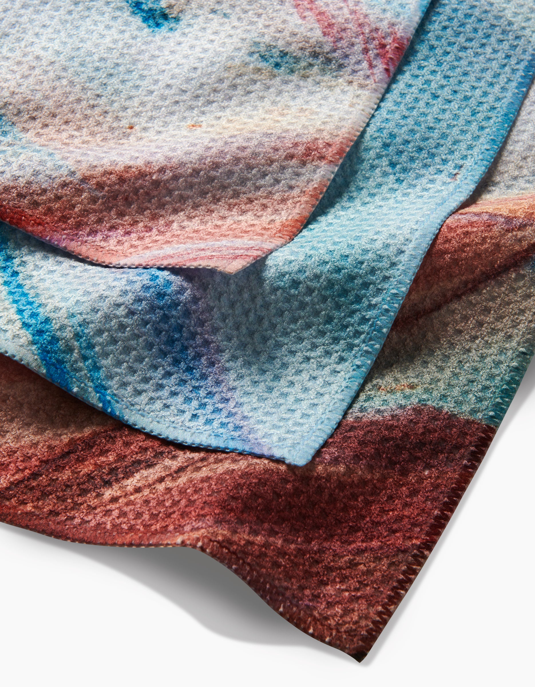 A close-up of two layered dishcloths, Golden Hour in the Rockies by Kelly Peters, featuring a waffle weave from recycled materials and abstract multicolored designs in blue, red, and brown on a white background.