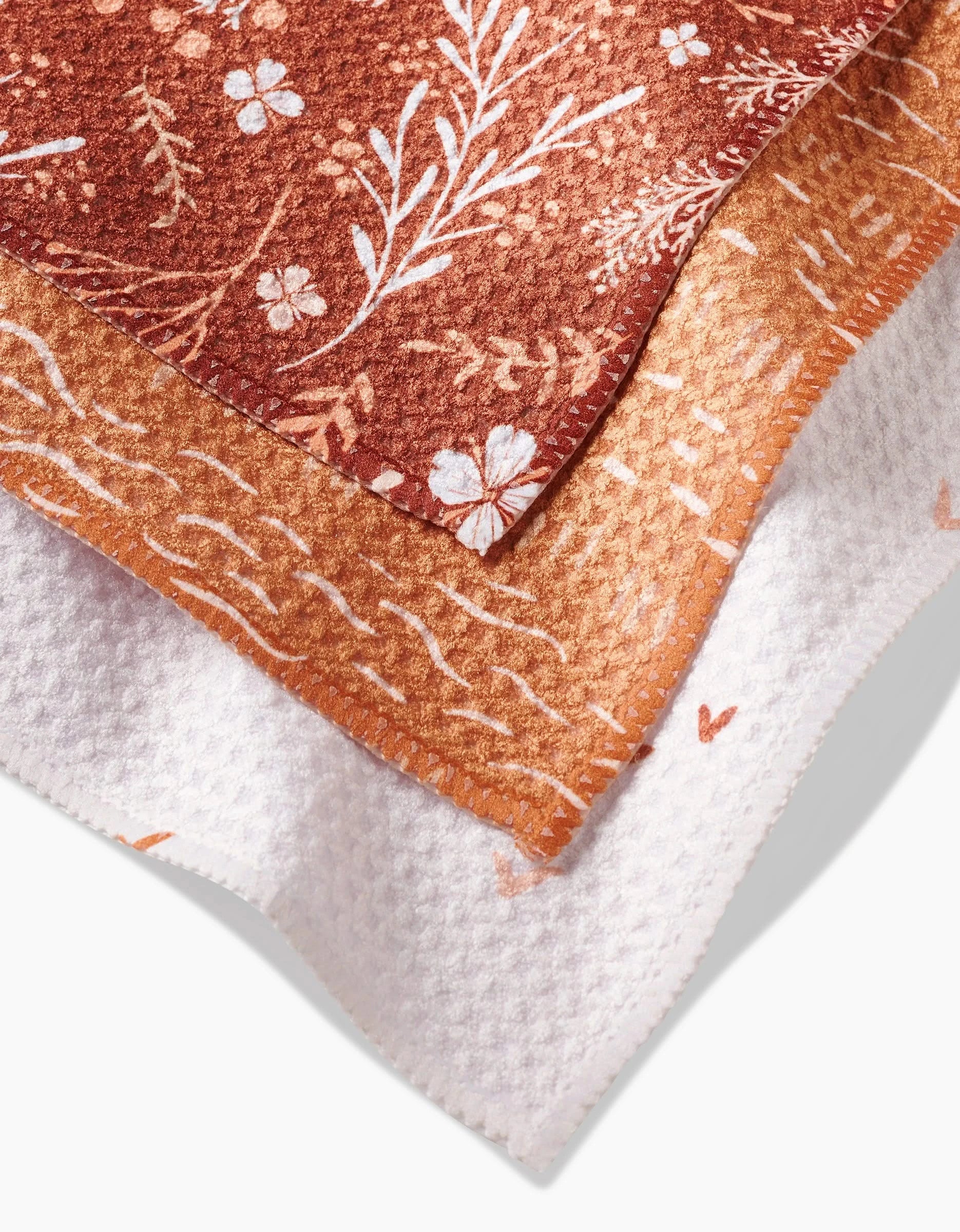 The Danelys Sidron Gust of Wind set includes three recycled dishcloths: rust with white florals, orange with a wavy white pattern, and white with light orange motifs, stacked together for stylish eco-friendly cleaning.