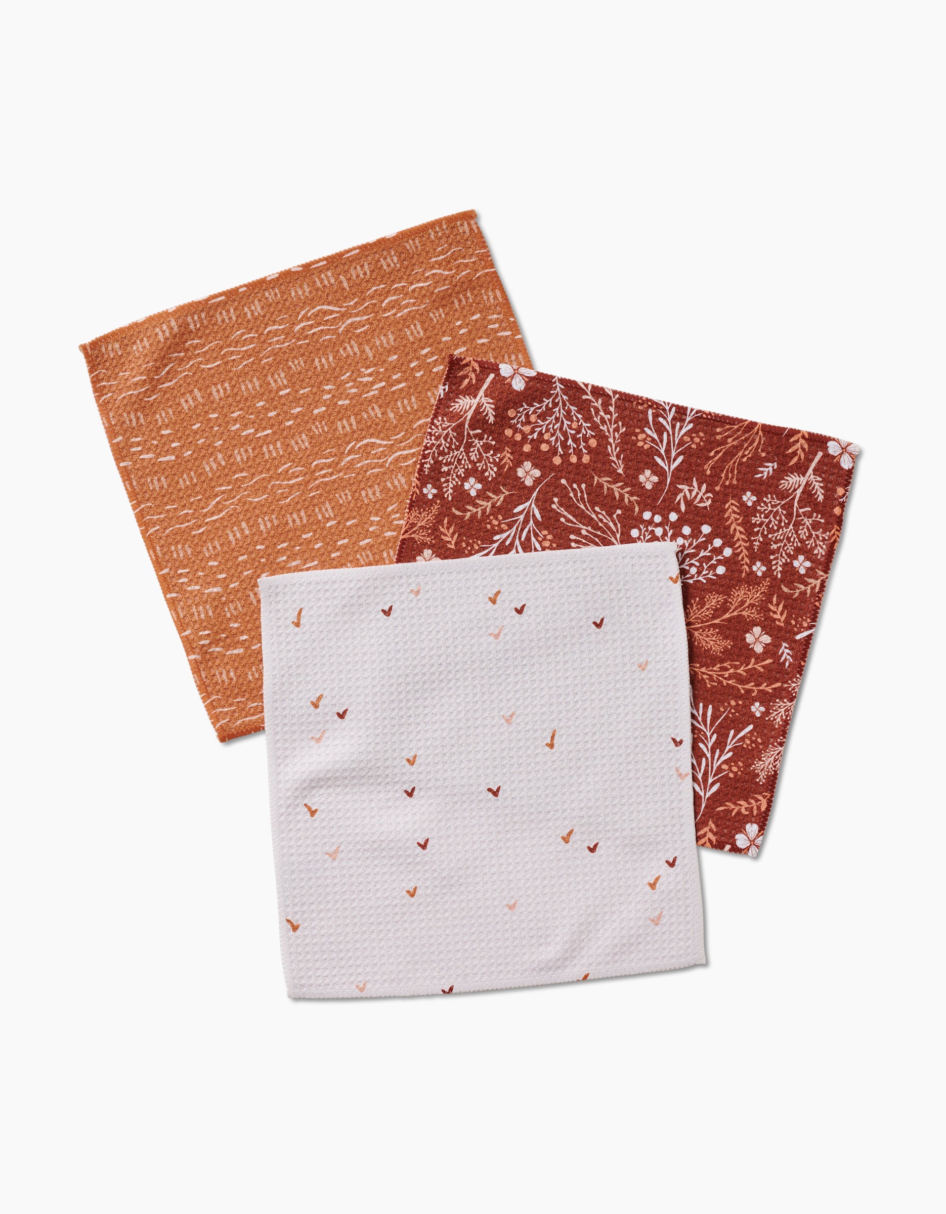 Three Gust of Wind cloth napkins by Danelys Sidron are shown: an orange geometric, a reddish-brown floral, and a super-absorbent white napkin with scattered heart shapes, all displayed on a white background.