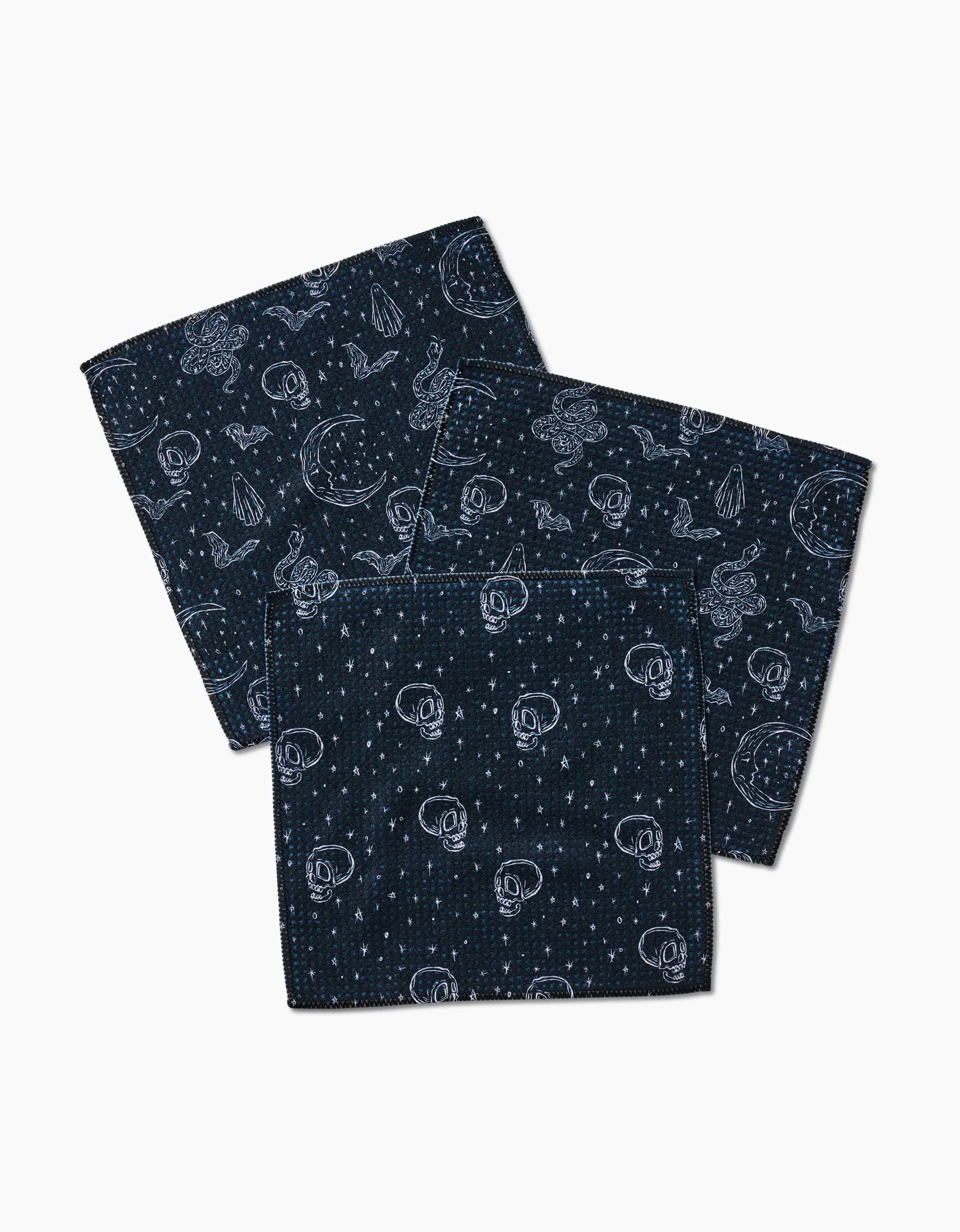 Aubrey Fairchild’s Halloween Hauntings set includes three navy blue cloth napkins made from recycled materials, featuring white-outlined skulls, stars, moons, and celestial Halloween designs on a white background.