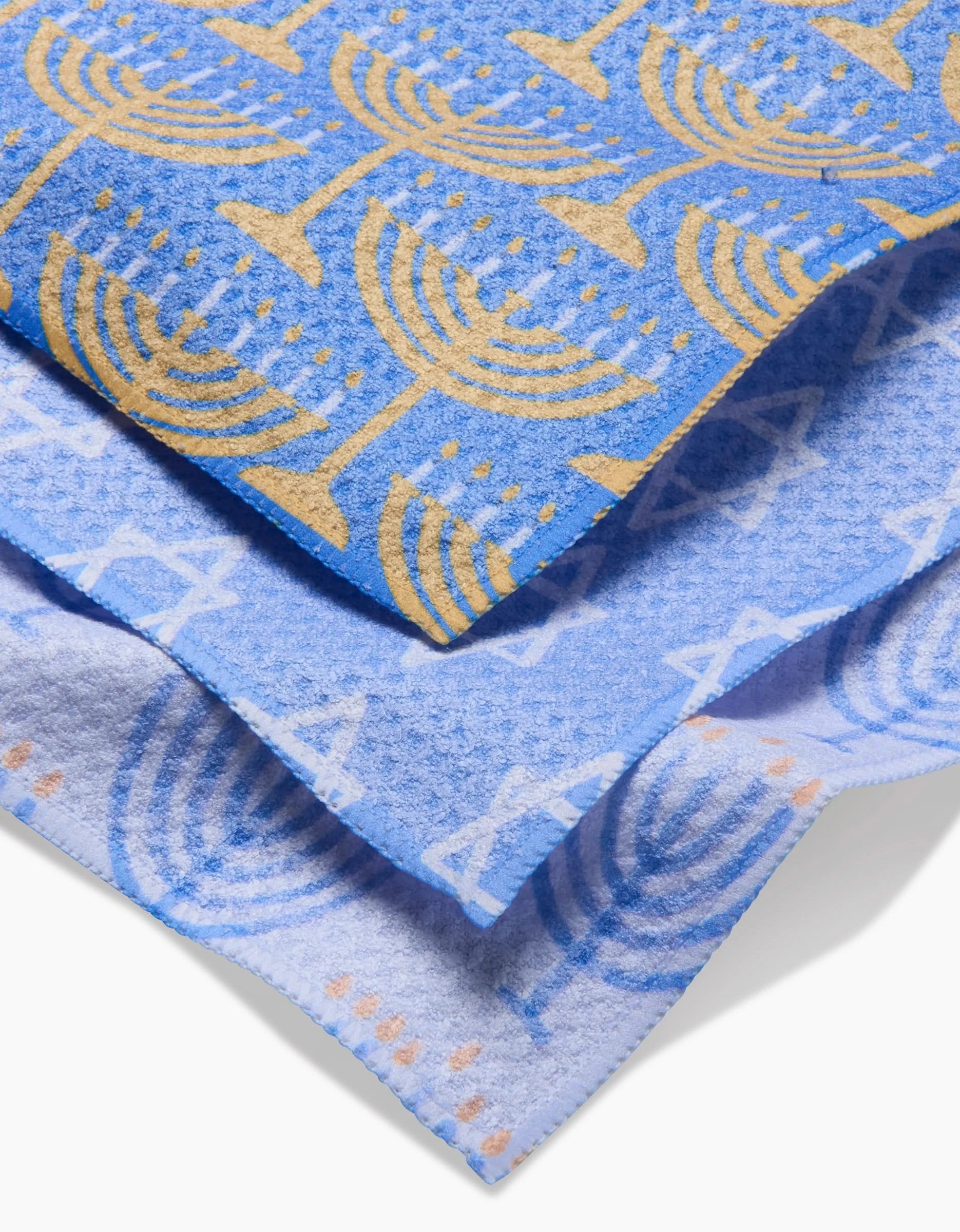 The Mindfulnice Hanukkah Lights set includes three blue and white tea towels stacked: yellow menorahs on top, blue menorahs in the middle, and blue Stars of David on the bottom. Made from absorbent Geoweave fabric.