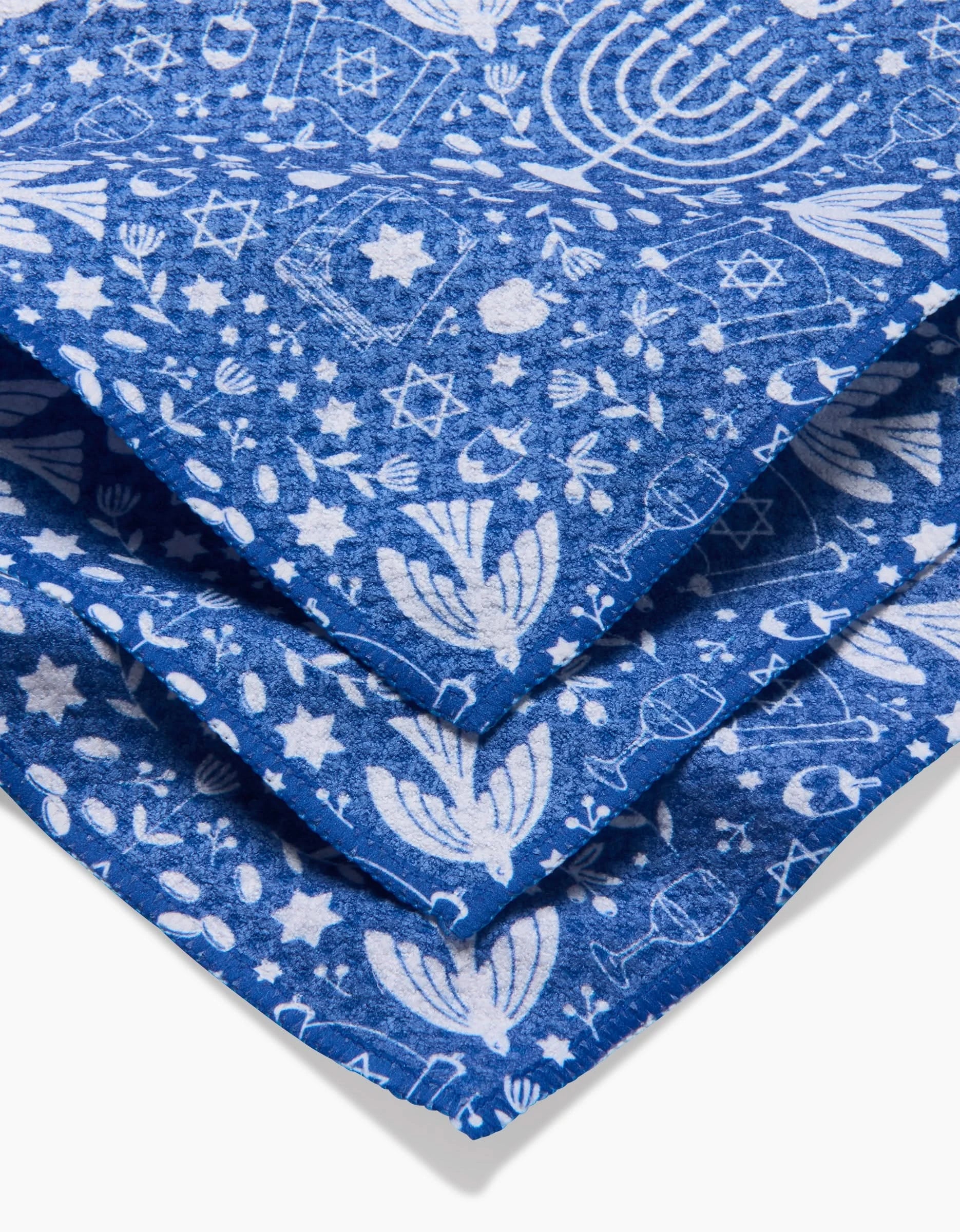 The Hanukkah Sameach kitchen tea towel by Megan Kampa features folded blue Geoweave fabric with white menorahs, Stars of David, dreidels, and festive Hanukkah patterns—perfect for holiday celebrations.