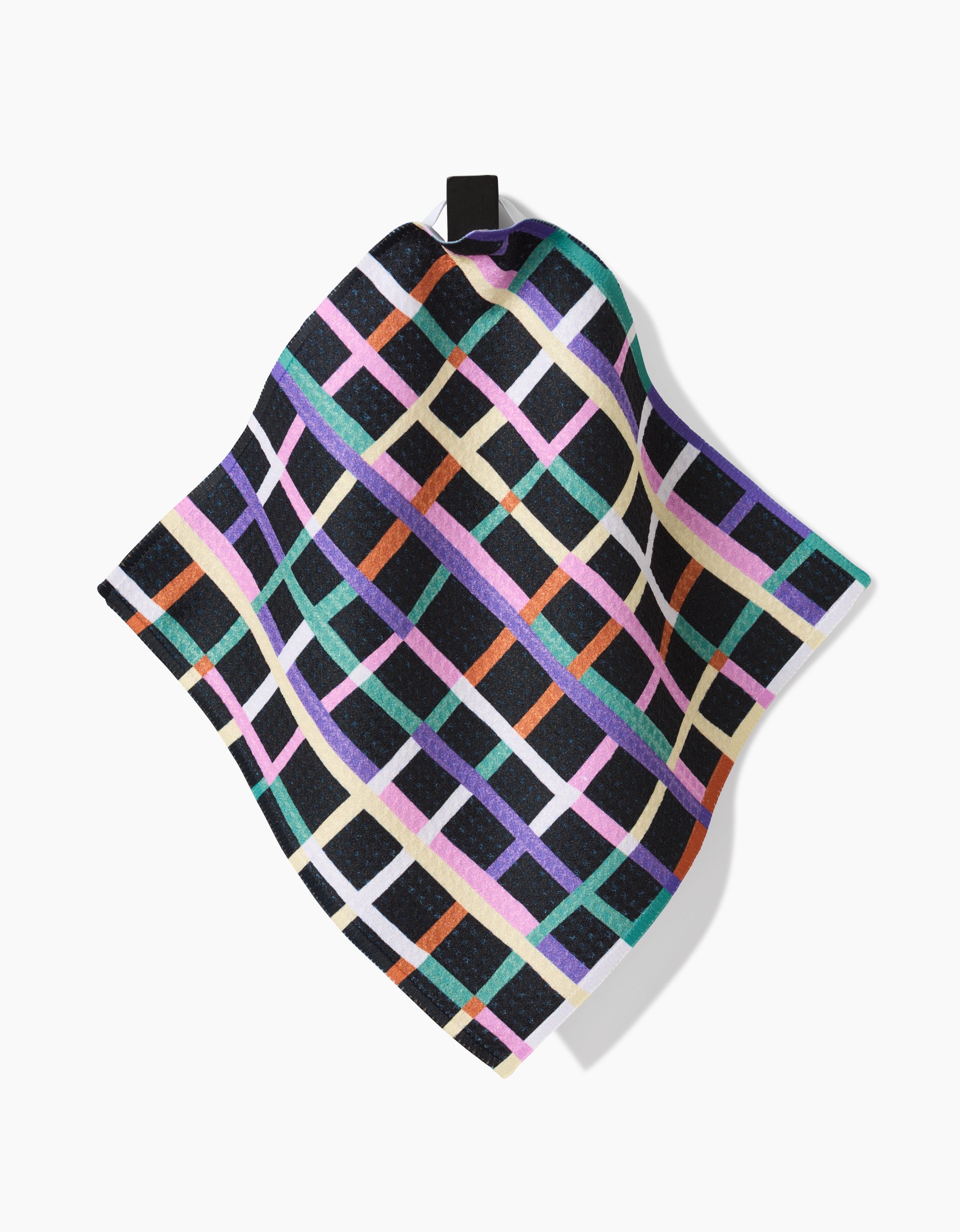 The Happy Plaid dish towel by Claire Iglesias features a checkered pattern in pink, orange, green, purple, and cream on black. Made from recycled materials, it hangs from a black loop against a white background.