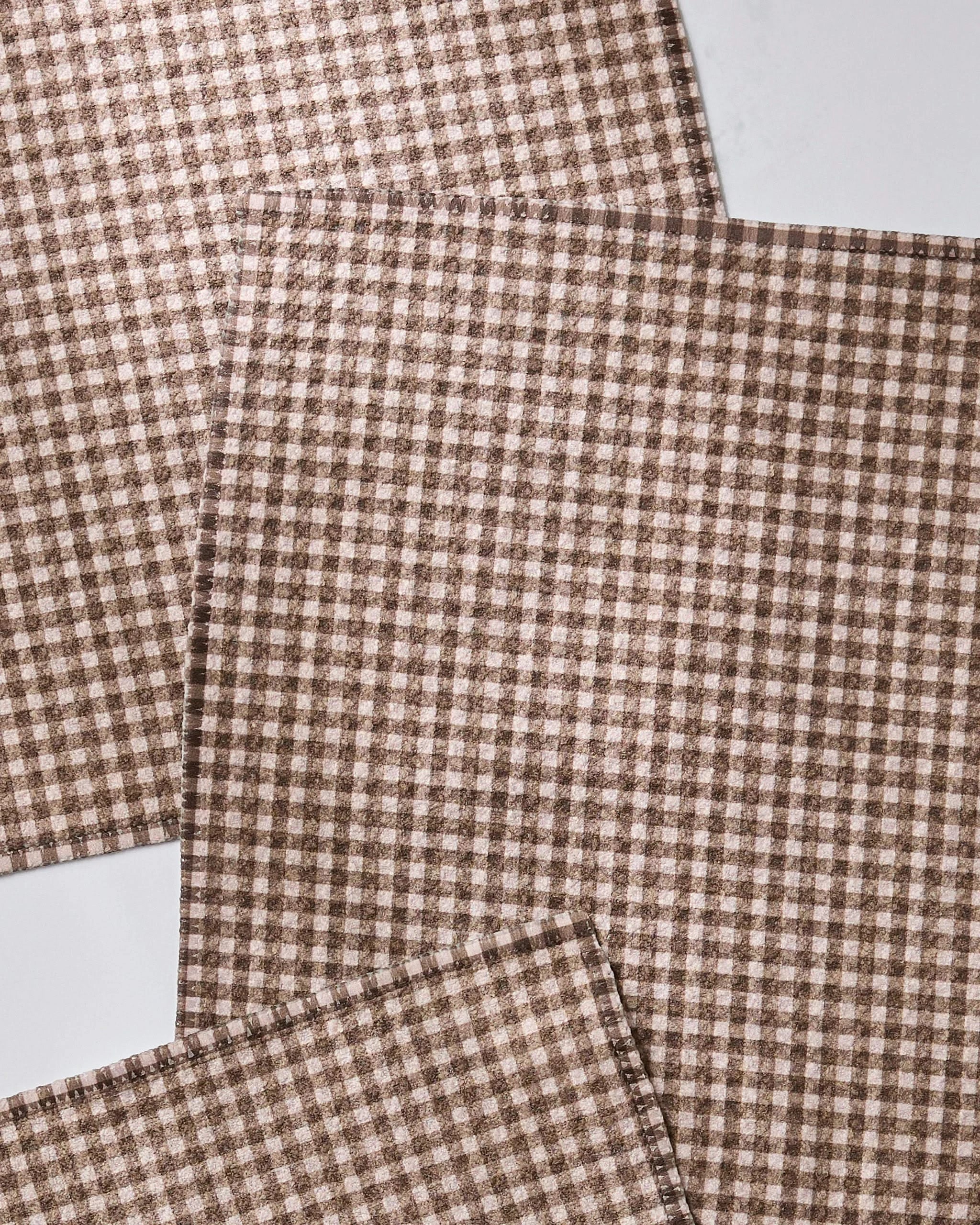 Three brown and white checkered Hearth napkins from Geometry’s super-absorbent dishcloth set are overlapped and laid flat on a white surface.