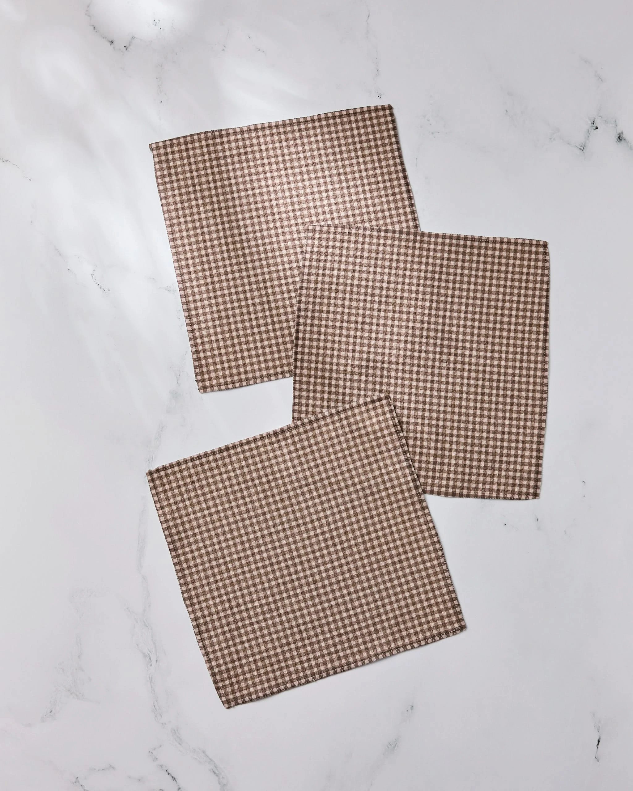 Three Hearth recycled dishcloths by Geometry, featuring a brown and white houndstooth pattern, are neatly arranged on a white marble surface, with two overlapping at the corners.