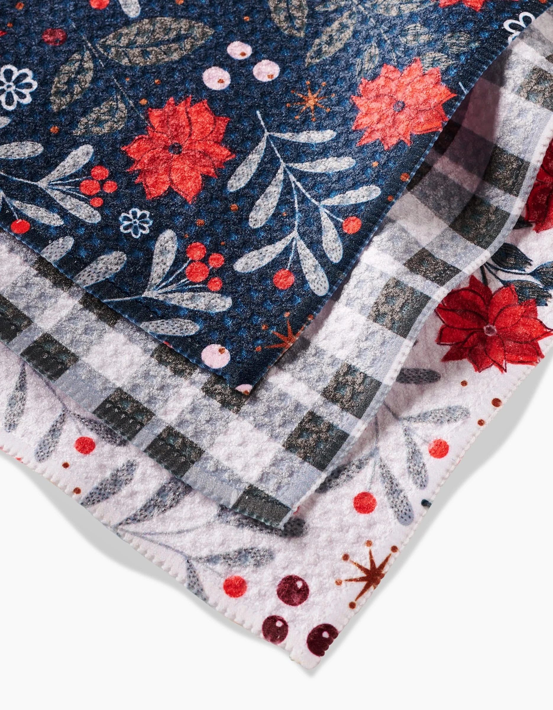 Close-up of two overlapping Charlie Rowan Designs Holiday Floral dishcloths: one with red flowers and green leaves on dark, the other with red flowers and a gray checkered pattern on white. Both are made from recycled materials.