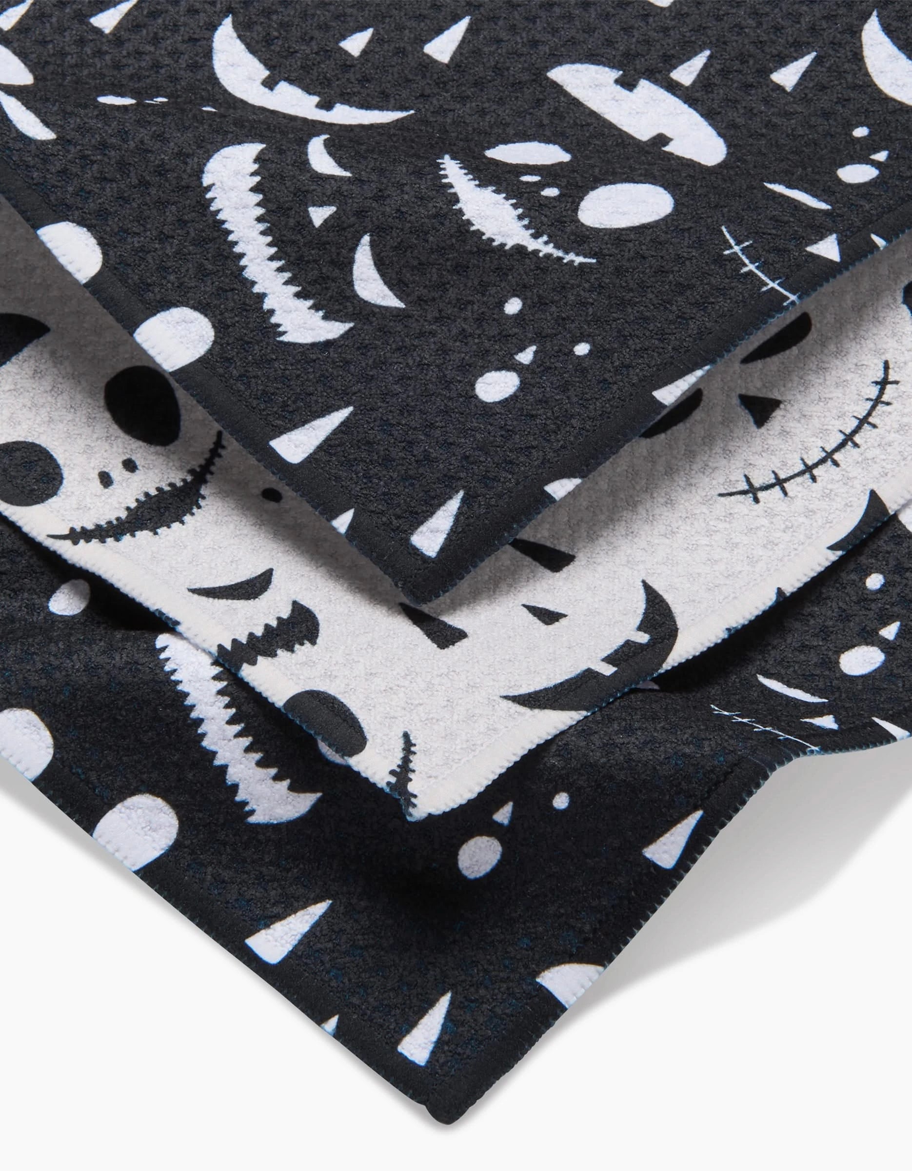 The Jack O Lantern Jamboree dishcloth set by Danelys Sidron includes three overlapping textured cloths made from recycled materials, featuring playful white Halloween faces on a black background—perfect for festive cleaning!.