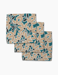 Three folded Janine napkins by Amy MacCready, made of Geoweave fabric, display a bold floral pattern in tan, black, and white on a teal background, stacked with each napkin partially overlapping the one beneath.