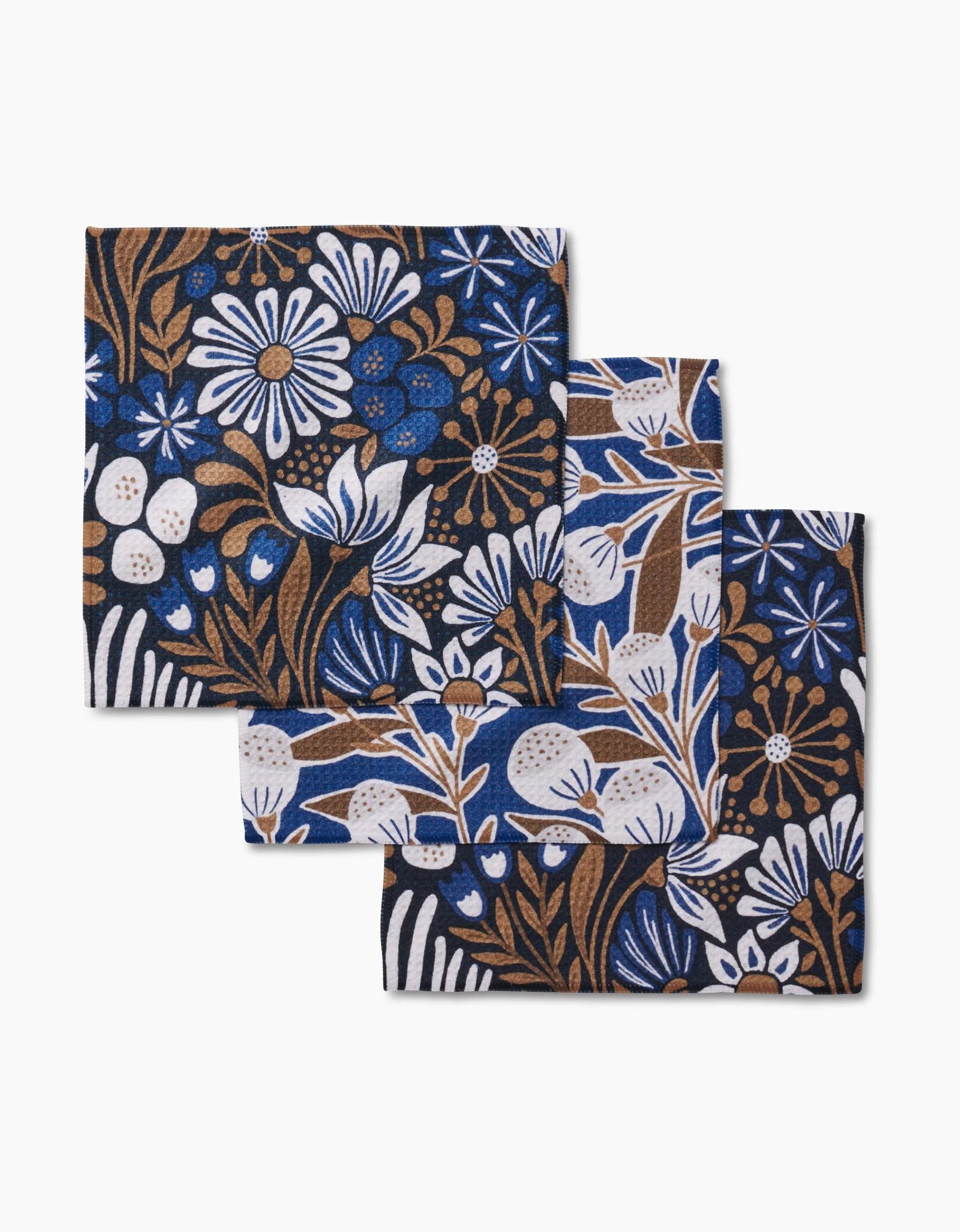 Three folded Leslie Flowers cloth napkins by Amy MacCready with a blue, white, and brown floral pattern are arranged overlapping on a plain white background.