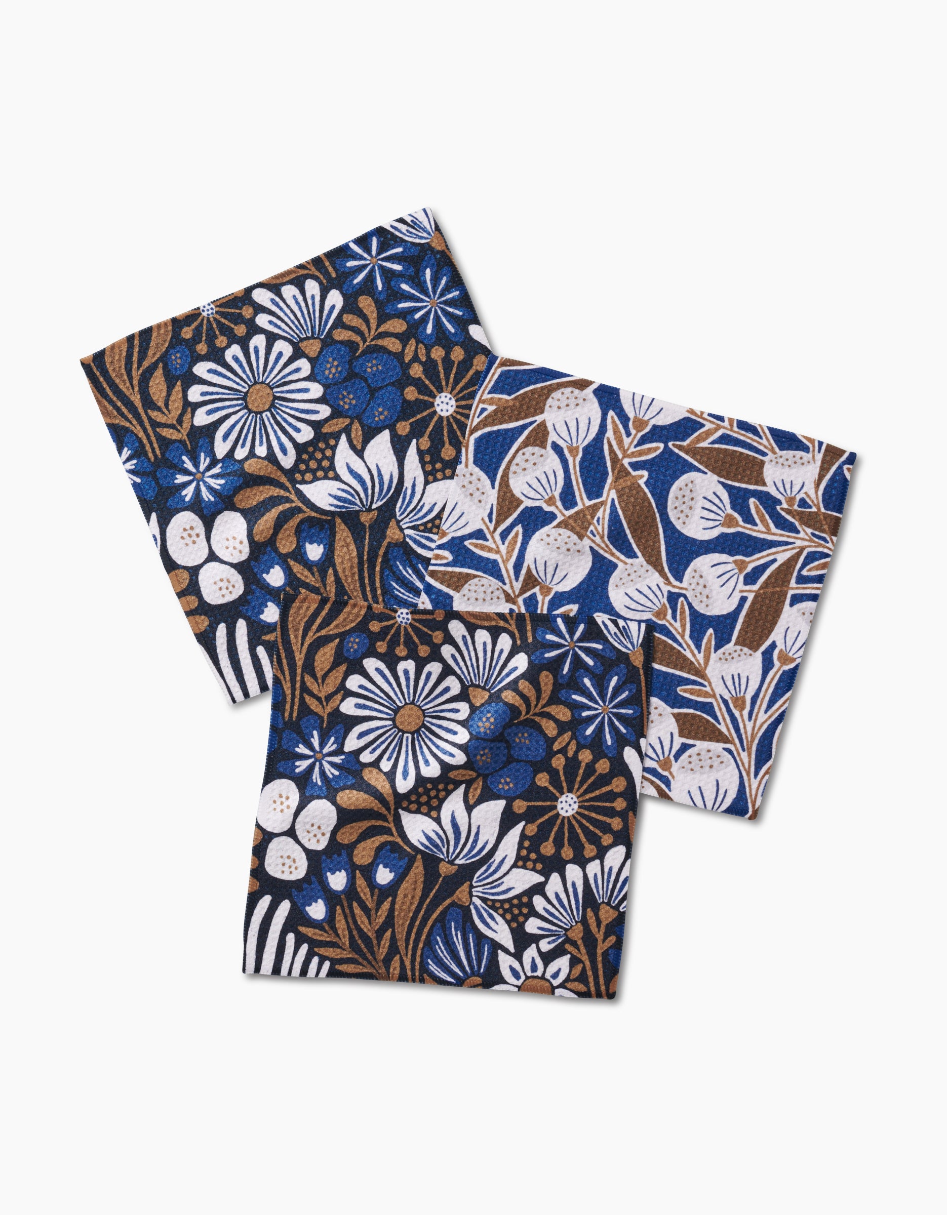 Three square Leslie Flowers napkins by Amy MacCready, featuring super-absorbent material and floral and leaf patterns in blue, brown, and white, shown overlapping on a plain white background.