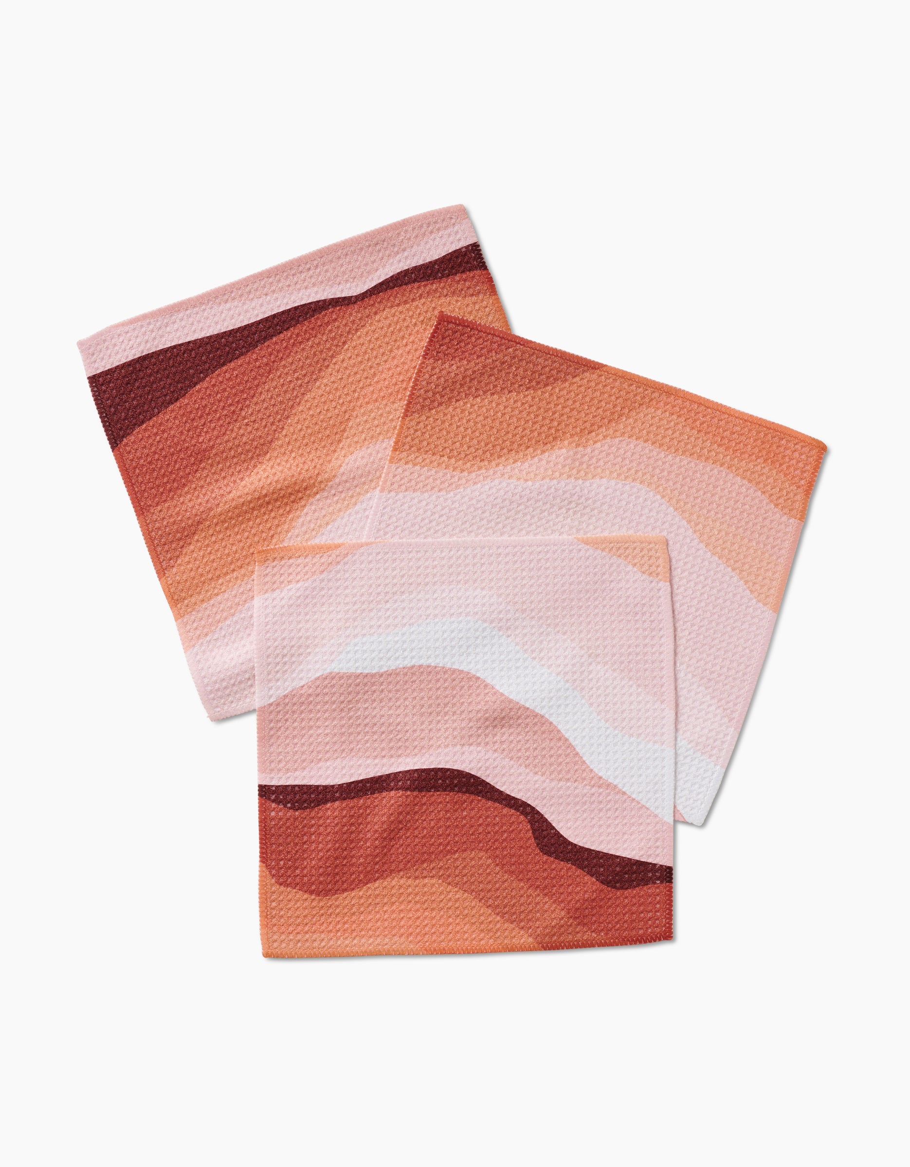 Three square GEOMETRY dishcloths from the Levels of Fall set, featuring abstract wavy patterns in pink, orange, and red Geoweave™ fabric, are arranged overlapping on a white background.