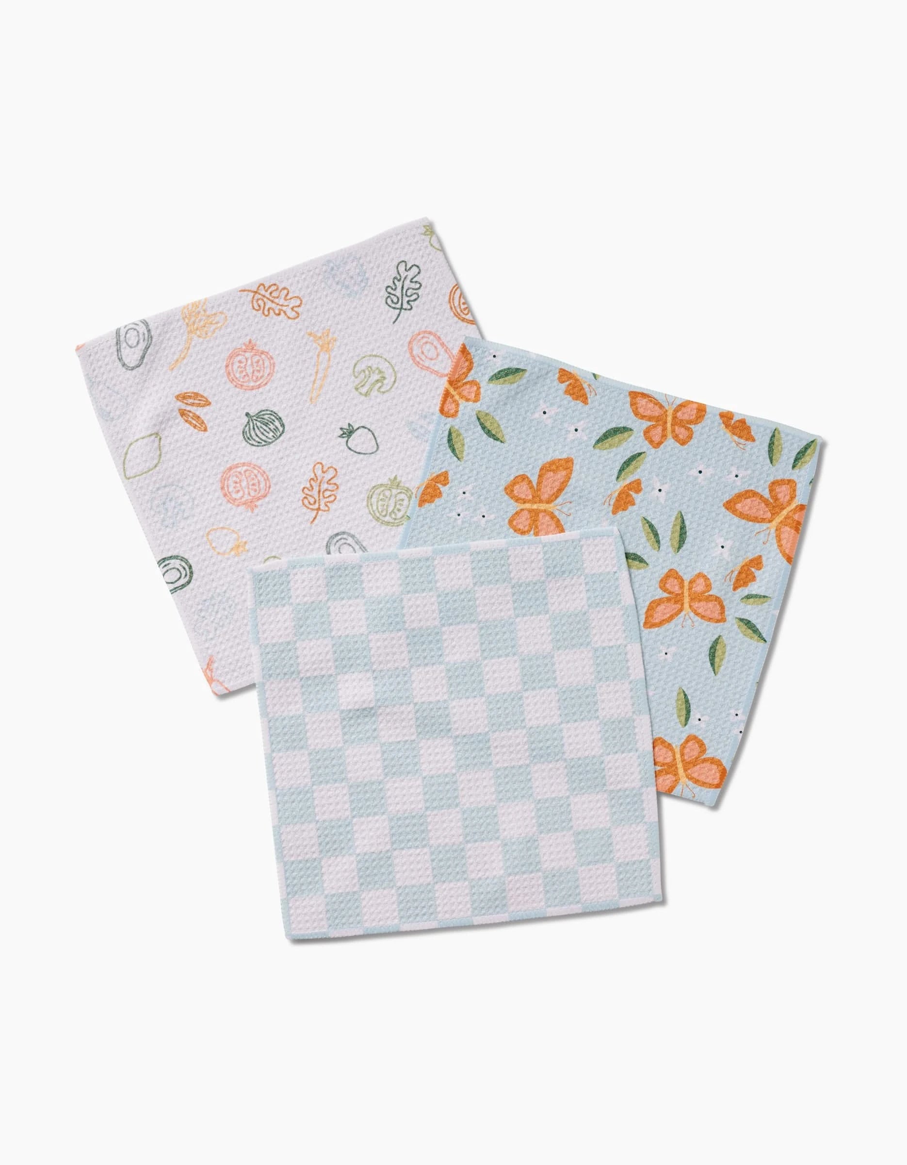 Three folded Life is Beautiful dishcloths by Sweet Simple Vegan overlap on a white surface: one has a pastel sushi pattern, one features orange flowers with green leaves on blue, and one displays a light blue and white checkered design.
