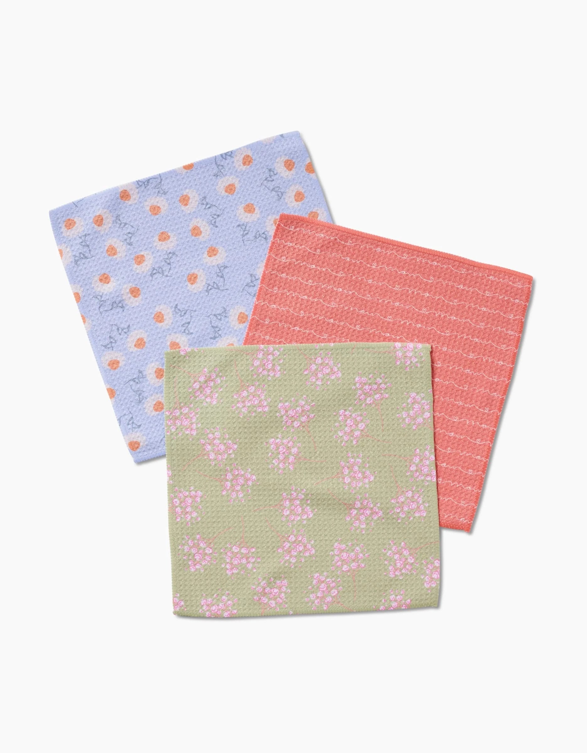 The Love of Mom dishcloth set by Sarah Reed features three square cloths—blue with orange dots and light blue shapes, coral with white lines, and olive green with pink and white flowers—overlapping on a white background.