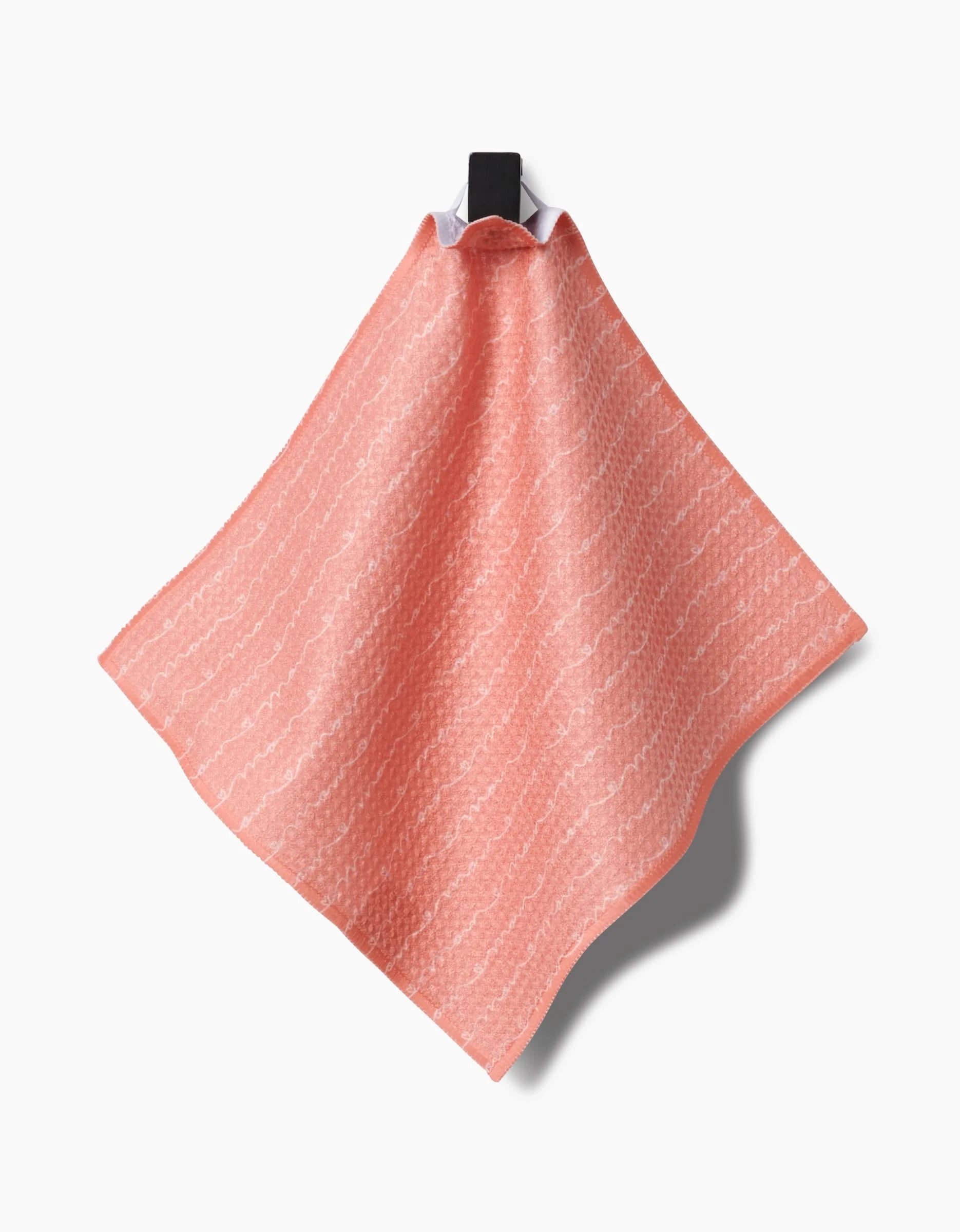 The Sarah Reed Love of Mom coral pink cloth, made from recycled materials and featuring a subtle white wavy pattern, hangs on a black hook against a white background with its shadow cast to the right.