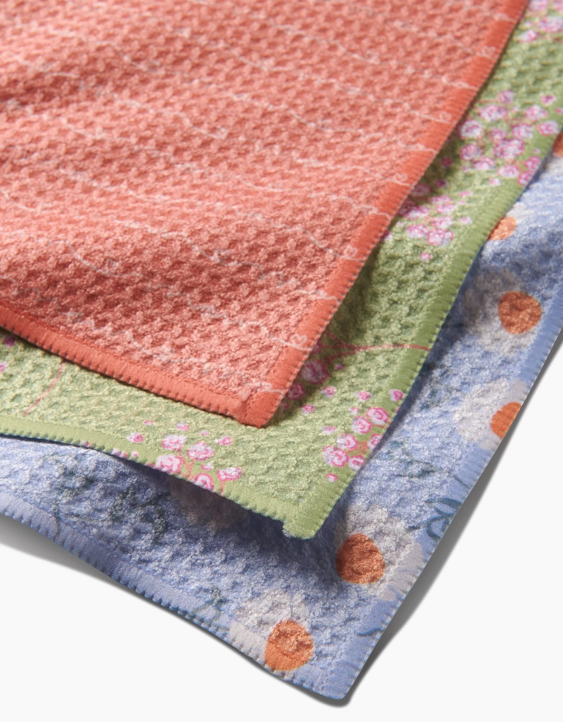 Three Love of Mom towels by Sarah Reed, made from recycled materials, are stacked: the top in coral with a floral pattern, the middle green with pink flowers, and the bottom blue with orange and white flowers.