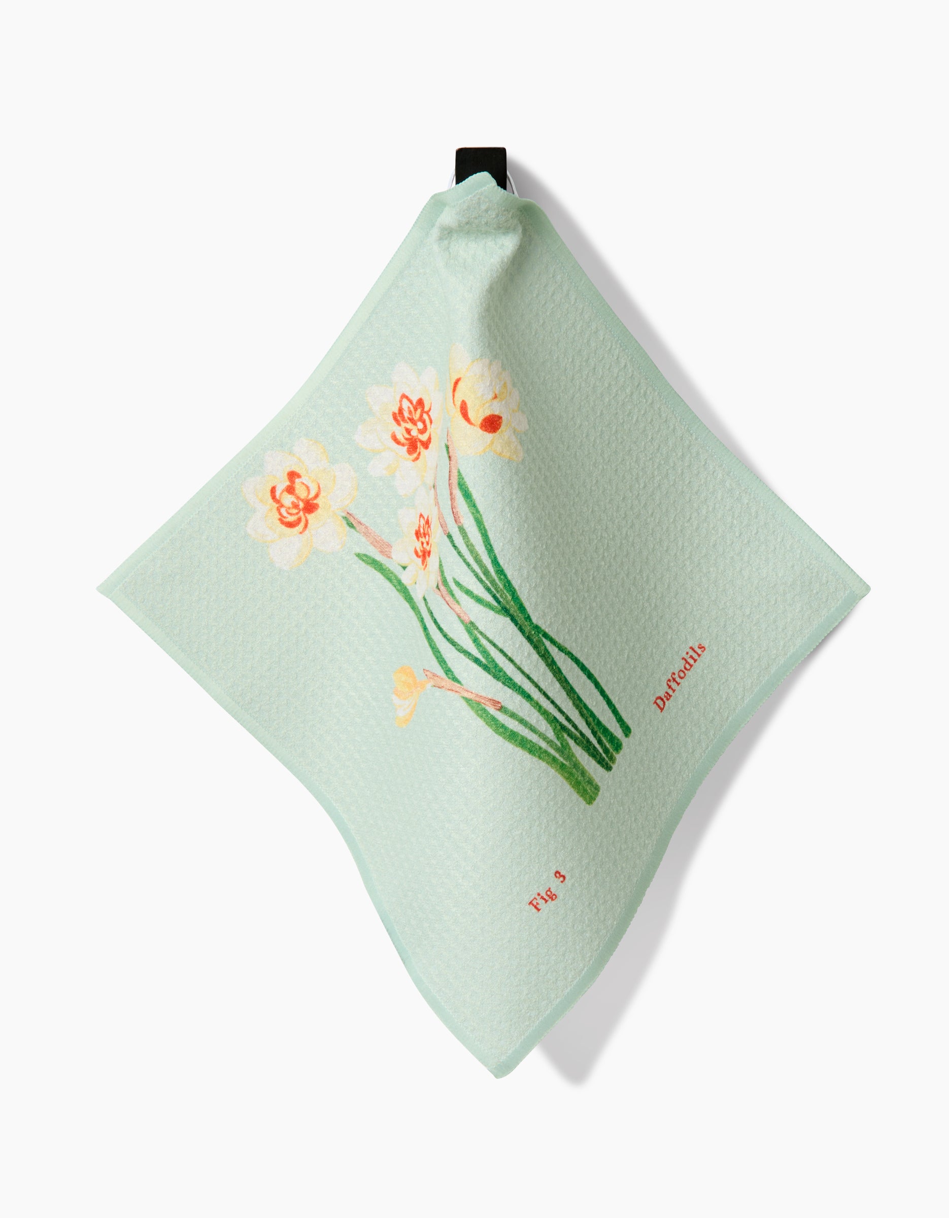 The March Daffodils by Linda Fox is a light green textured Geoweave fabric cloth featuring daffodil flowers, green stems, and This is Daffodil in red, with a black hanging loop at the top corner.
