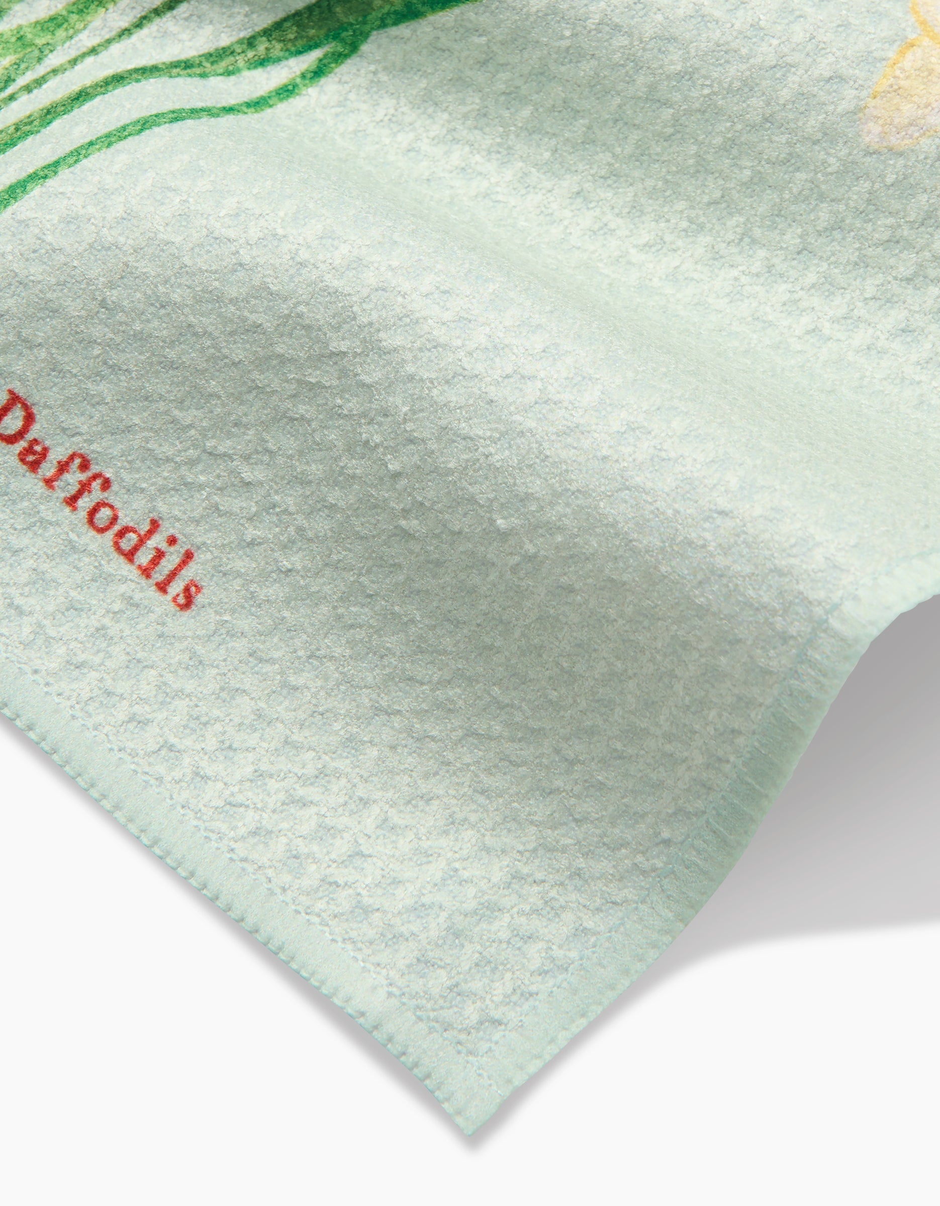 A close-up of Linda Foxs March Daffodils dishcloth shows its textured light blue Geoweave fabric, the word Daffodils in red, and part of a yellow daffodil illustration at the top edge.
