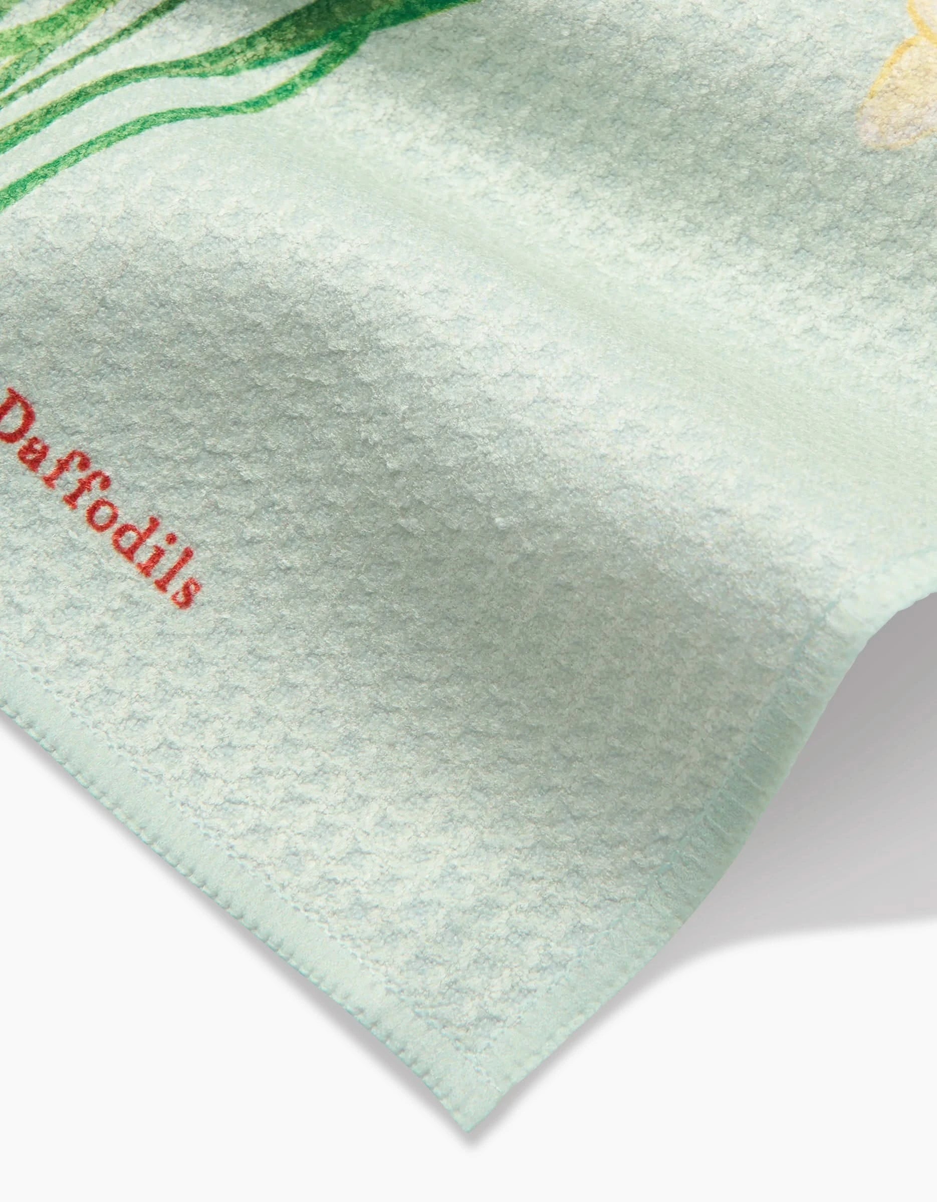 A close-up of Linda Foxs March Daffodils dishcloth shows its textured light blue Geoweave fabric, the word Daffodils in red, and part of a yellow daffodil illustration at the top edge.