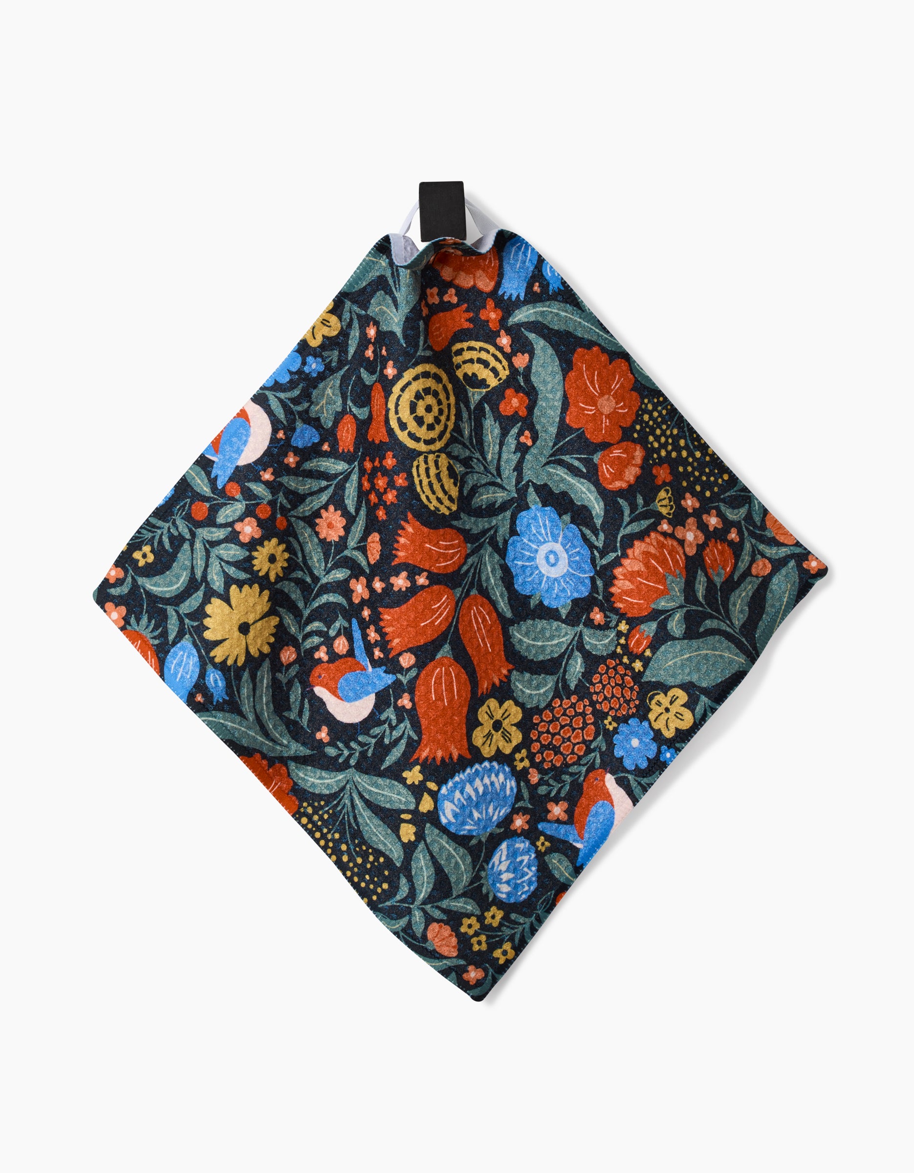 The Marsh Wren by Danelys Sidron is a square napkin made from recycled materials, featuring a vibrant floral and leaf pattern in red, blue, yellow, and green on dark fabric, with a small black fabric loop at one corner.