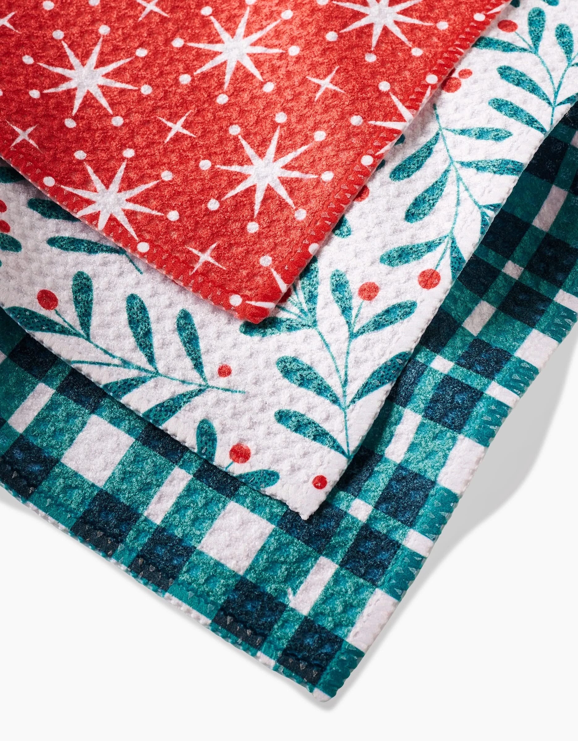The Merry Berry set by Charlie Rowan Designs includes three stacked dishcloths in red with white stars and dots, white with teal leaves and red berries, and teal-blue plaid—all crafted from durable Geoweave fabric.