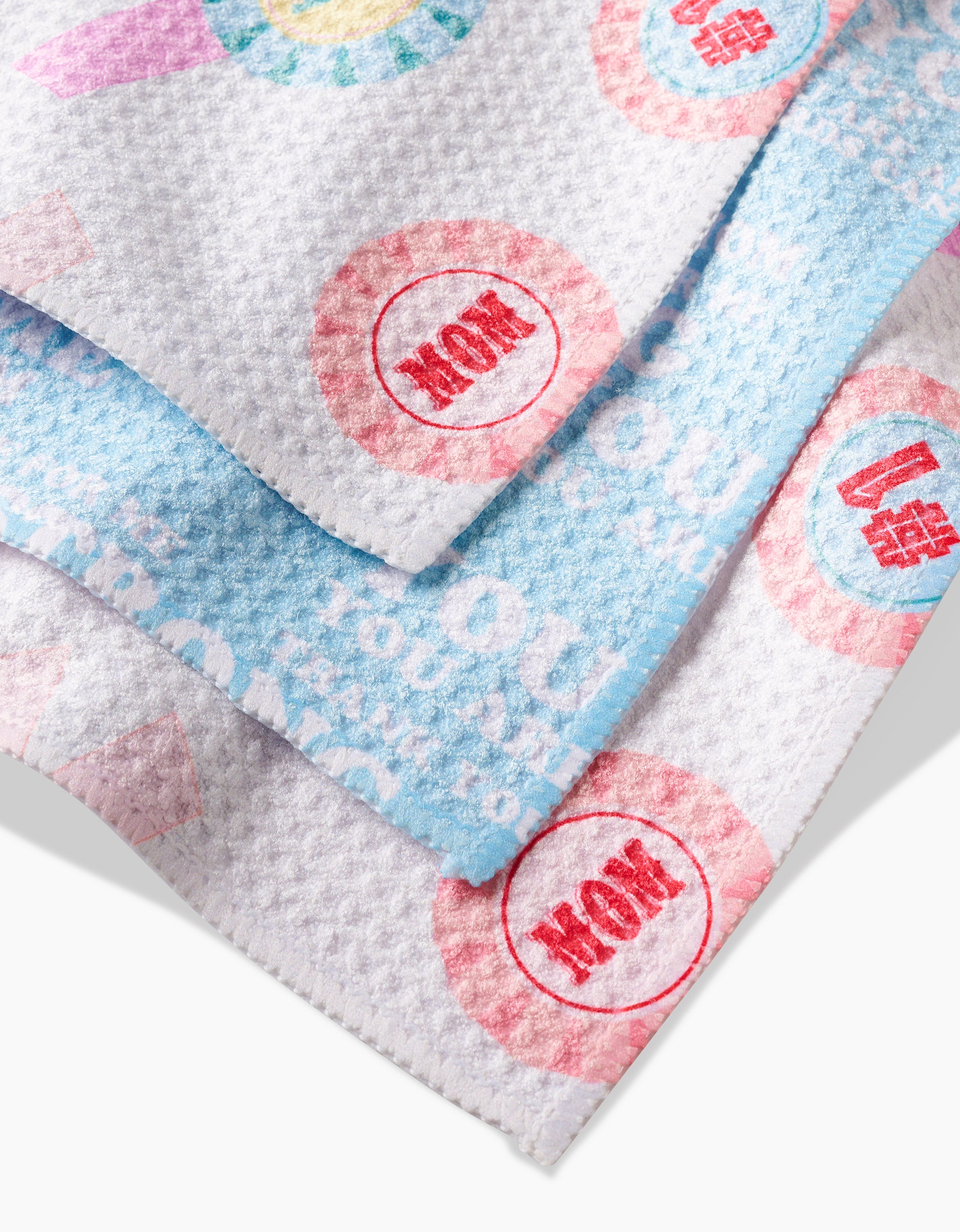 Three GEOMETRY Mom Mantra dishcloths, crafted from recycled materials, are layered together. Mainly white with blue and pink accents, each features faded Mom Mantra text and red WOW stamps on colorful patterns.