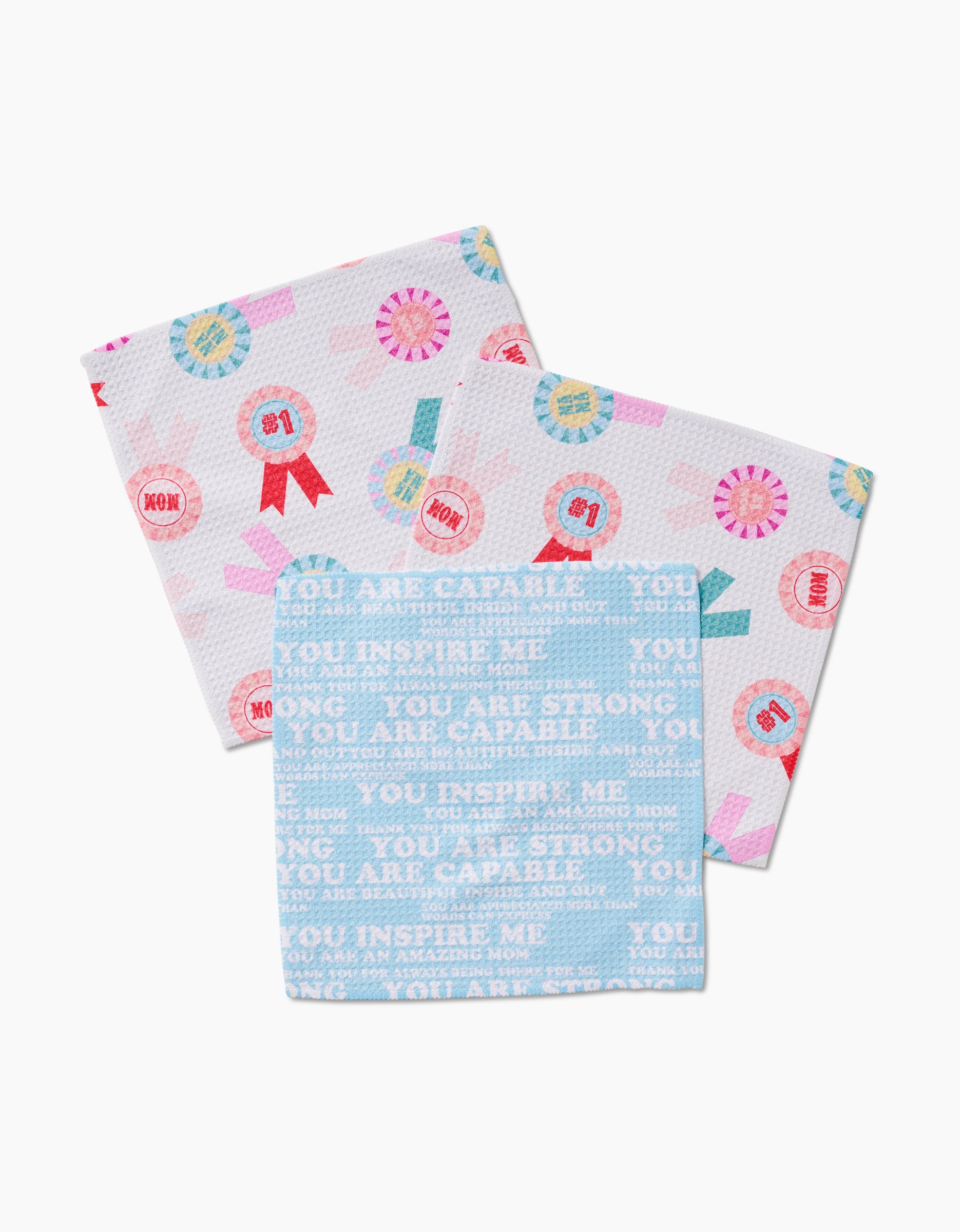 The GEOMETRY Mom Mantra set includes three patterned Geoweave dishcloths: two with trophy ribbons reading #1 Mom in pink, blue, and red, and one with light blue affirmations like “You are capable” and “You inspire me.”.