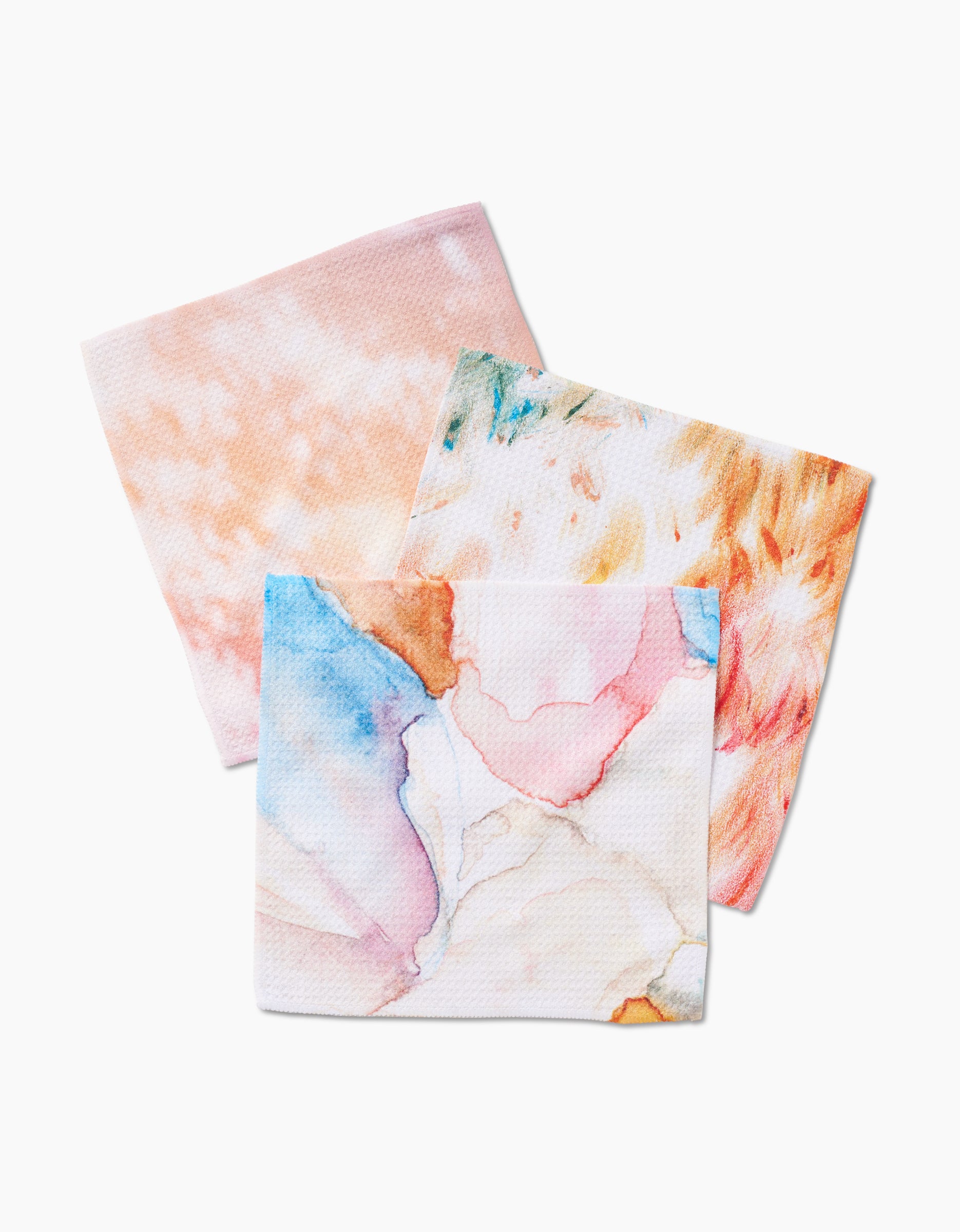 Three GEOMETRY Nice Water square cloths made from Geoweave fabric—one peach, one orange and green, and one blue, pink, and beige—feature watercolor patterns and are arranged overlapping on a white background.