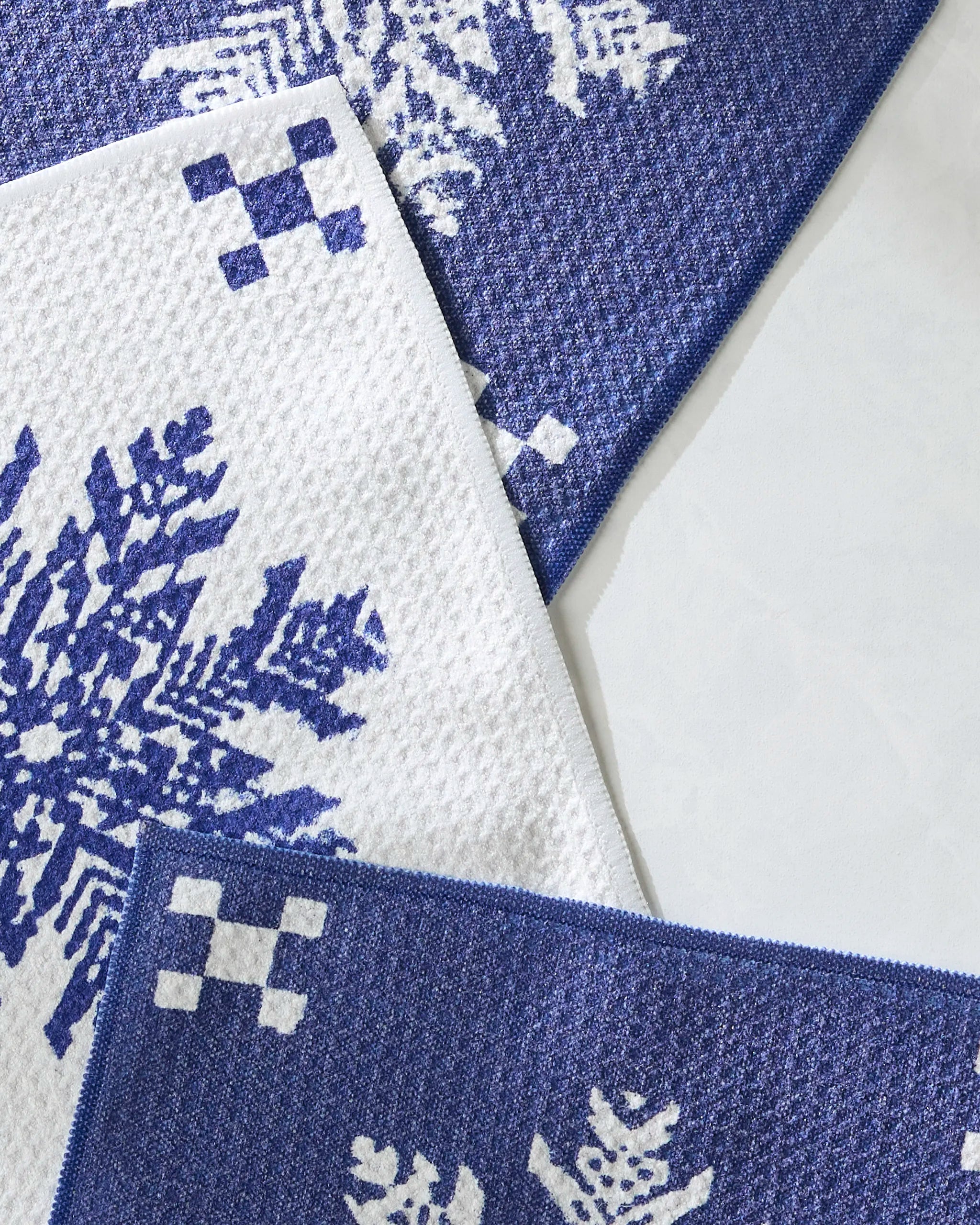 Nieve towels by Geometry are blue and white with overlapping large knit snowflake and checkered corner patterns on a light gray background, crafted from recycled materials.