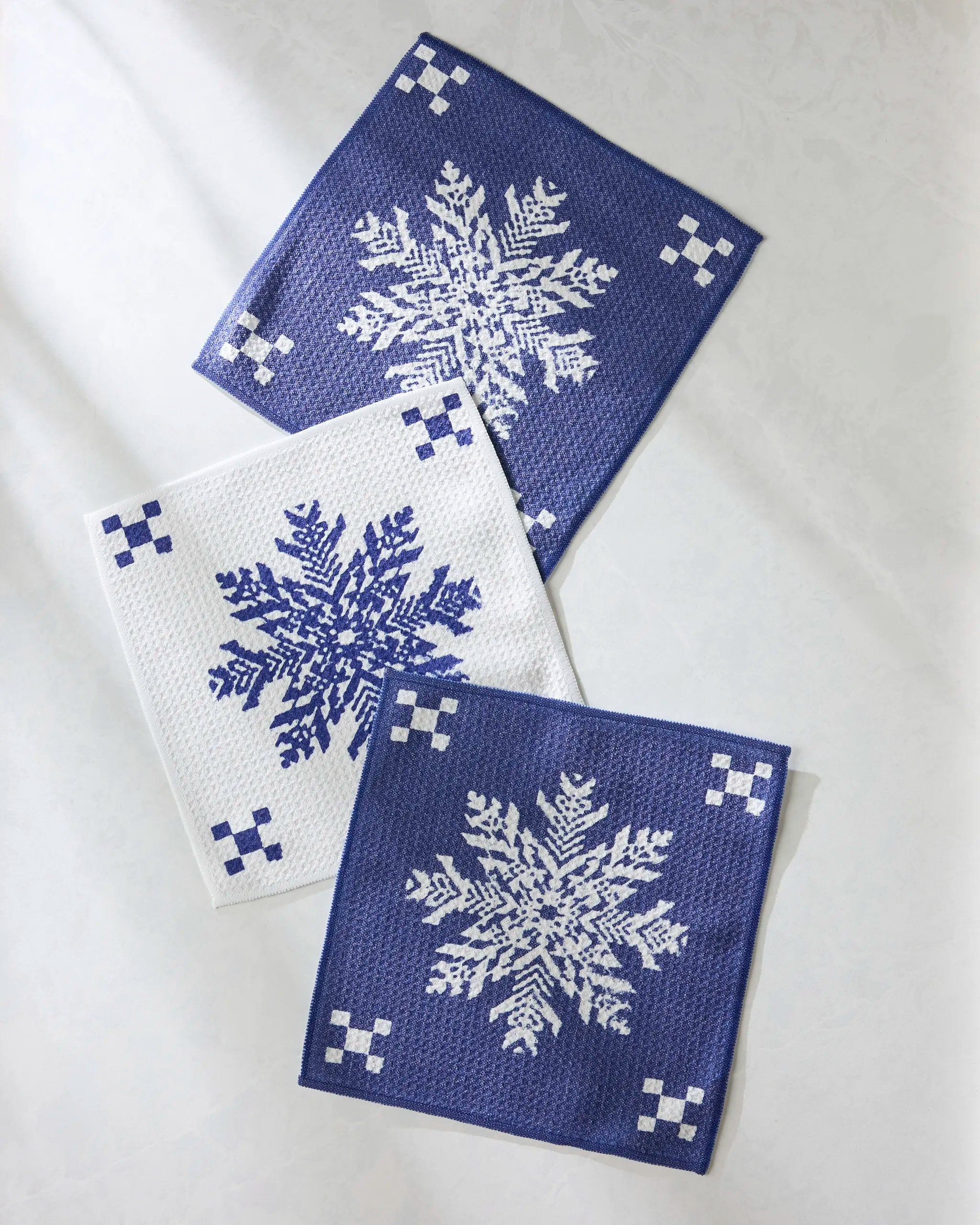 The Geometry "Nieve" set features three recycled cloths: two dark blue with white snowflake cross-stitching and one white with a blue pattern, arranged overlapping on a light surface for a charming, eco-friendly touch.