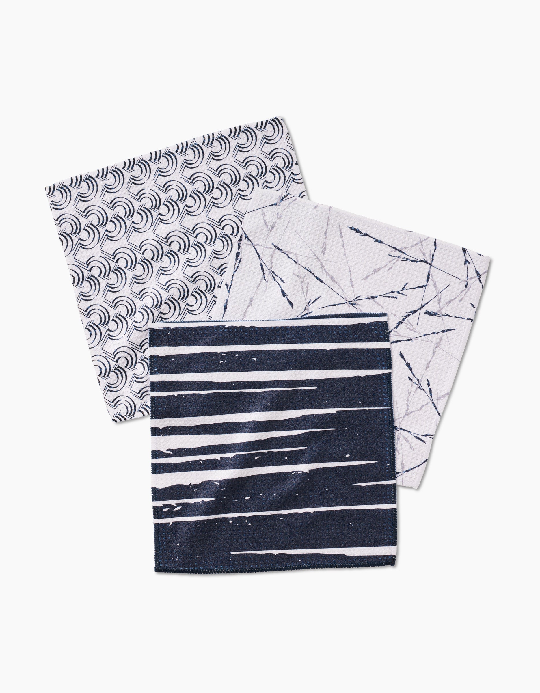 GEOMETRY’s Night Reeds set features three super-absorbent square dishcloths crafted from recycled materials—one white with navy waves, one with abstract lines, and one navy with stripes and spots—shown overlapping on a white background.