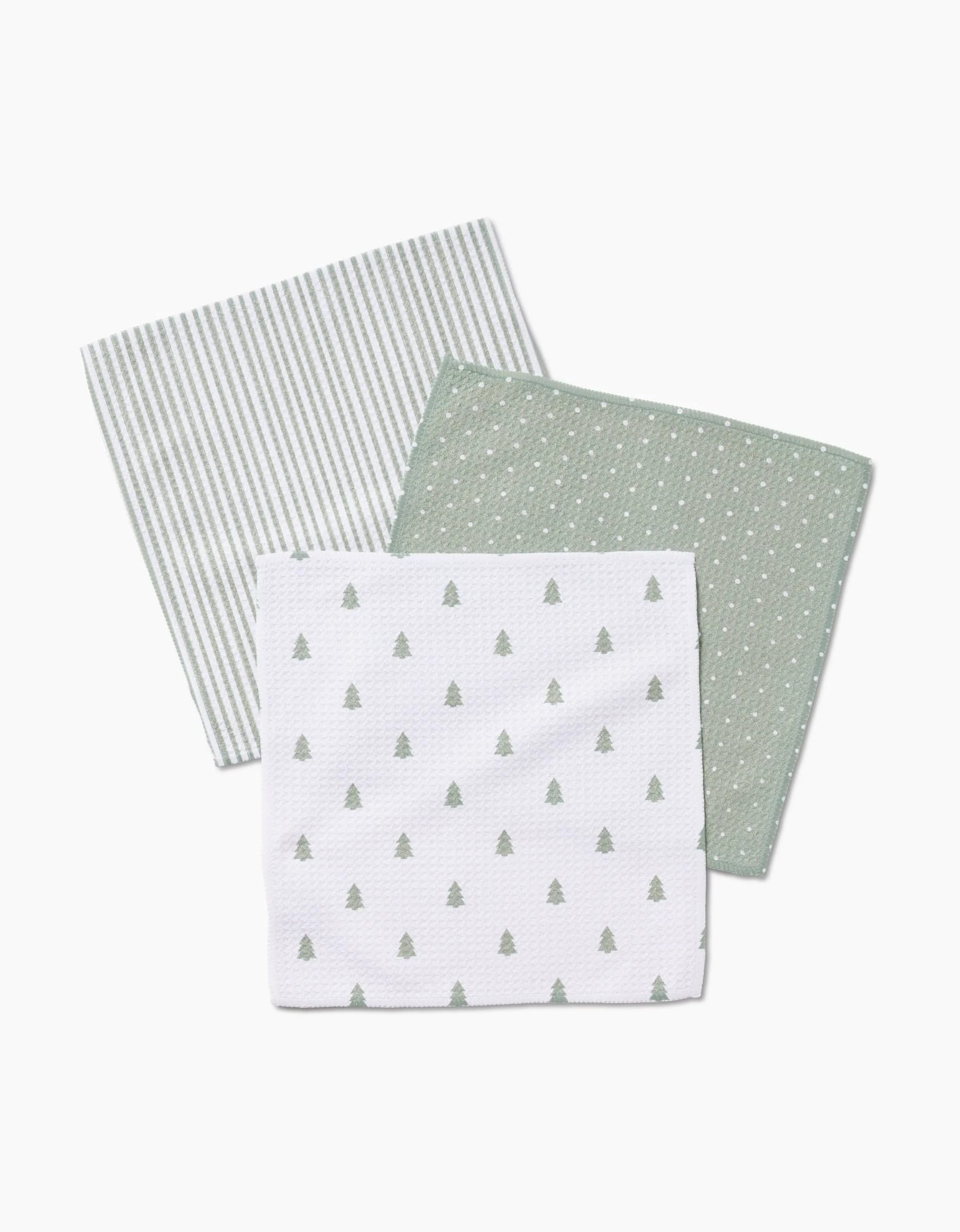 Three Noel cloth napkins by Kailee Wright, made from soft Geoweave fabric—one with grey stripes, one green with white dots, and one white with small green pine trees—are arranged overlapping on a white background.