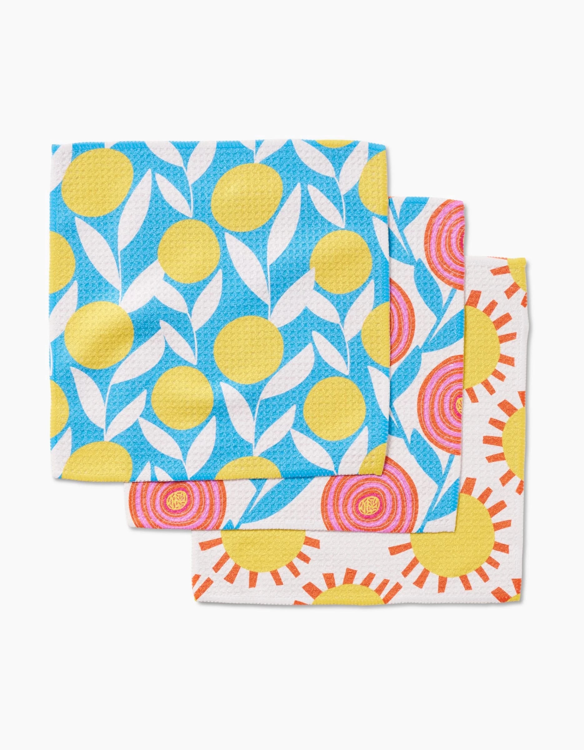 Three Parade dishcloths by Lizzie Clark are stacked, each made from Geoweave fabric and featuring vibrant sun-inspired designs in blue, yellow, pink, orange, and white. The top cloth highlights yellow circles and white leaves on blue.