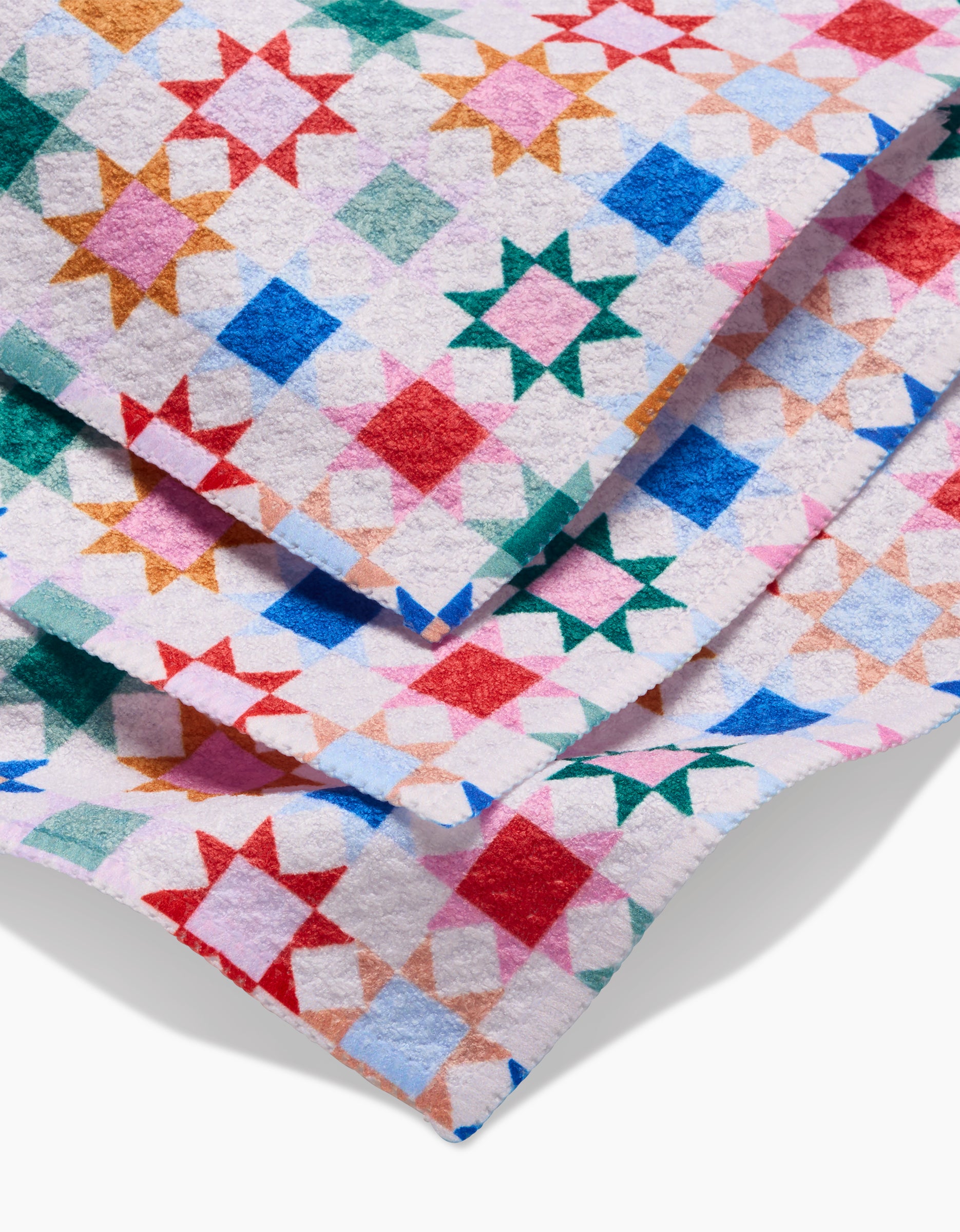 The Patchwork Picnic dishcloth by Brittany Jepsen features colorful Geoweave fabric with a geometric quilt pattern in red, green, blue, pink, orange, and white. Its shown folded to reveal its soft, textured surface and layered edges.