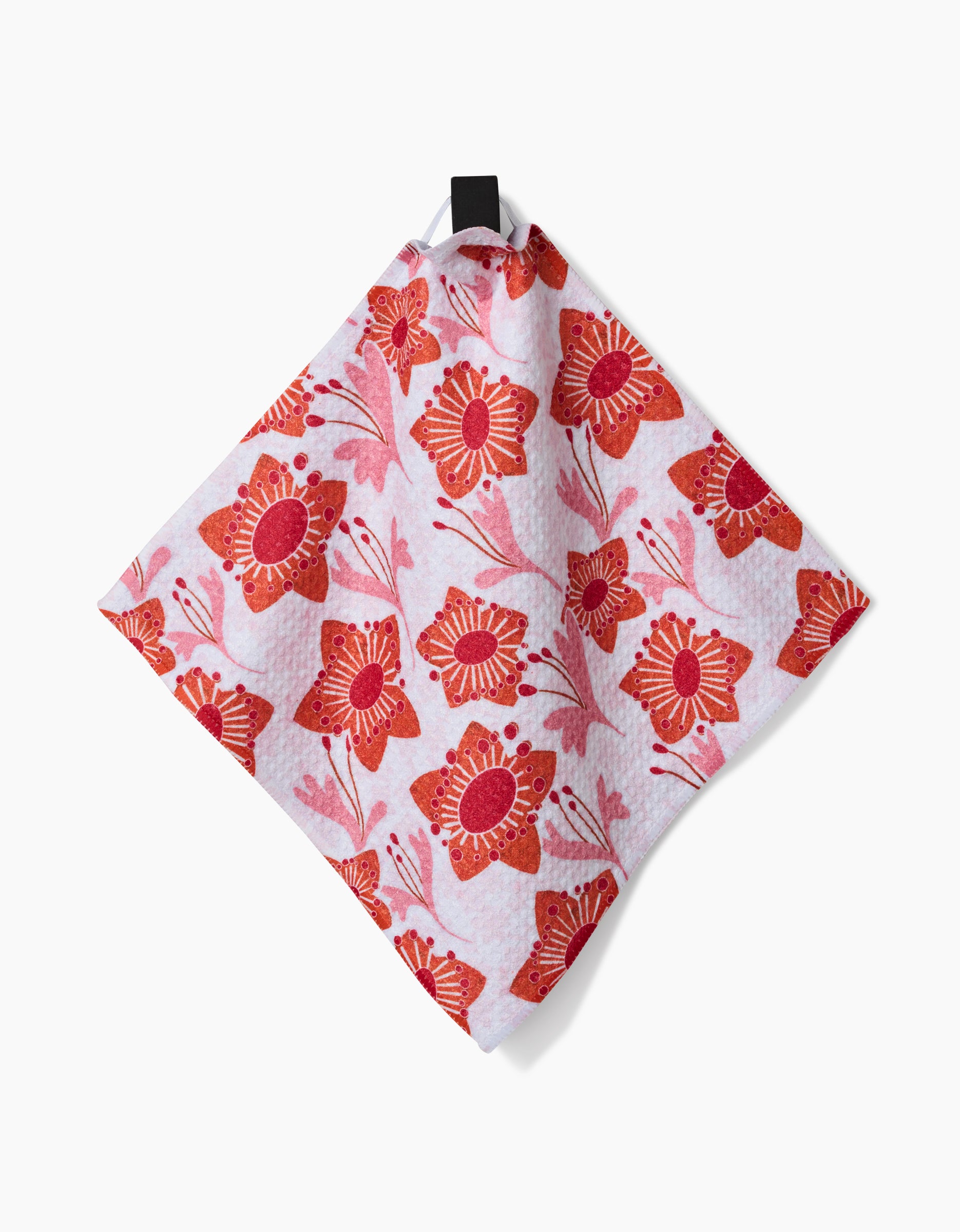 The Petal Paradise dishcloth by Whitney Urquhart, featuring large red and orange stylized flowers on pink Geoweave fabric, hangs from a black loop against a white background.