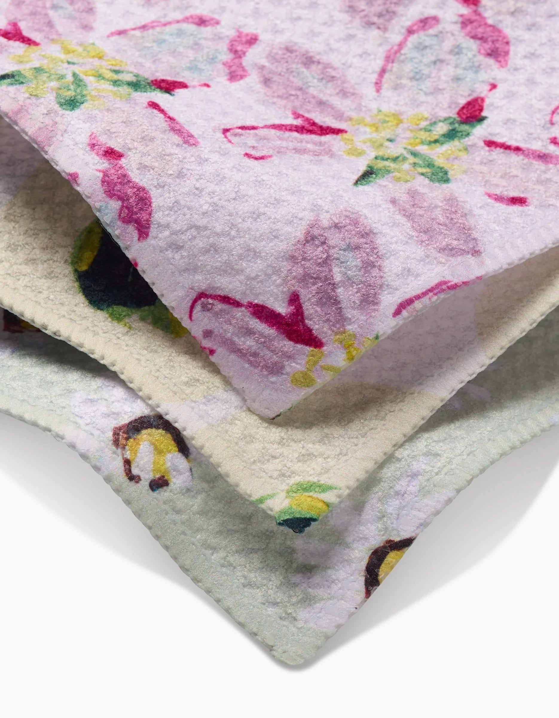 Stacked Petals dishcloths by Ania Zwara Art, each with unique floral patterns, are made from recycled materials—top features pink blooms, middle has yellow and purple flowers, and bottom is accented with multicolored designs.