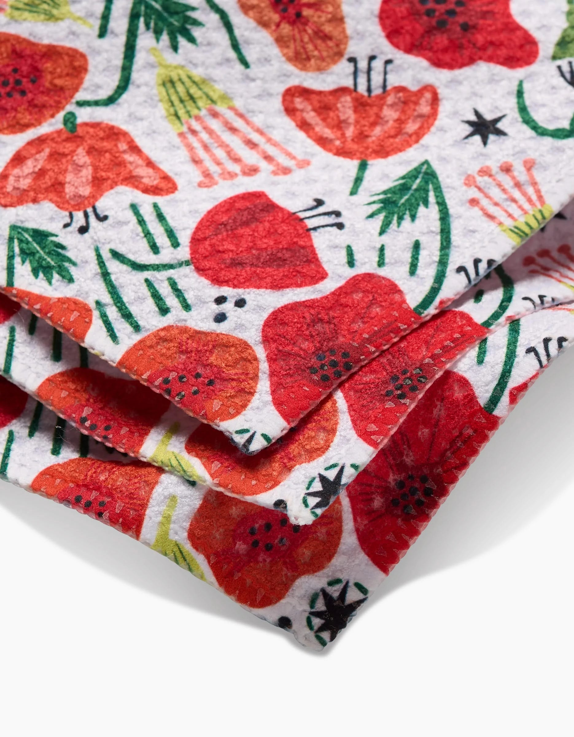 Close-up of Elisabeth Wing’s Poppy Power Geoweave fabric, showcasing vibrant red poppies with green stems and leaves on white. The folded layers highlight its versatile use as a dishcloth or for other household needs.