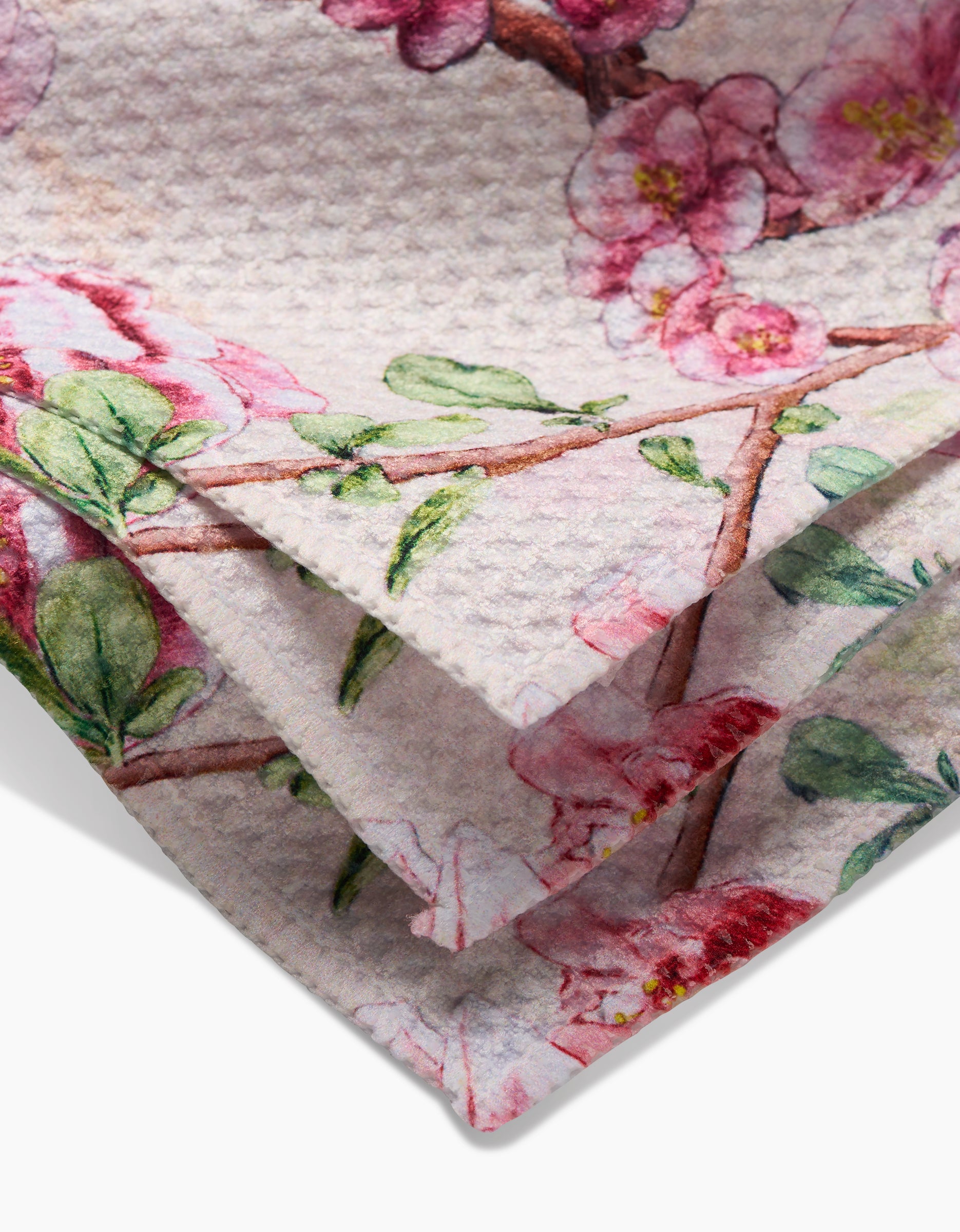 A close-up of folded Pascale Dilger Primroses Geoweave fabric dishcloths with a textured surface, printed with pink primroses and green leaves on branches against a light background.