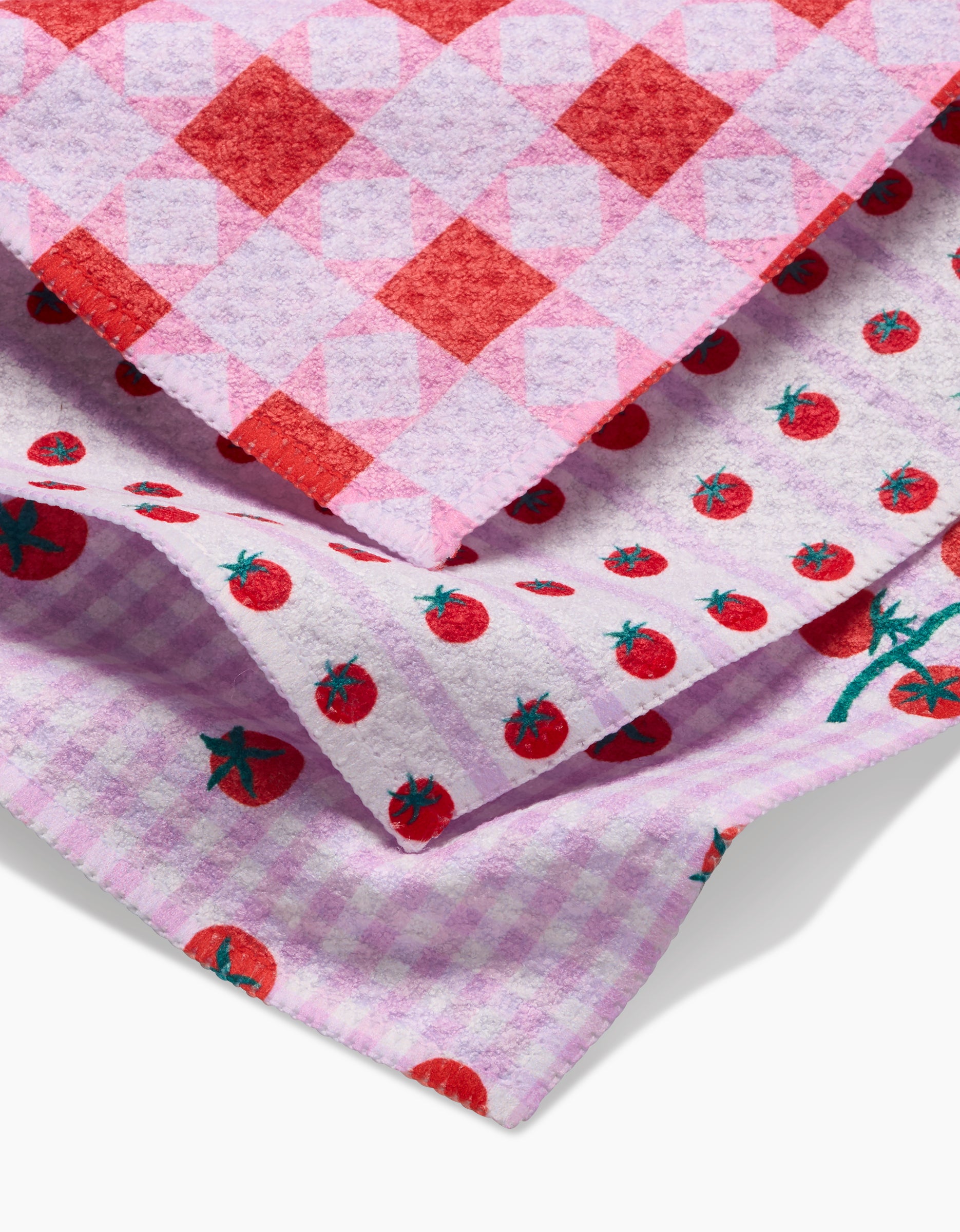 Three colorful dishcloths from Brittany Jepsen’s Quilt Tomato Harvest set overlap: the top features red and pink checks, the middle shows red tomatoes on white, and the bottom is light purple gingham with tomato motifs—all crafted from Geoweave fabric.