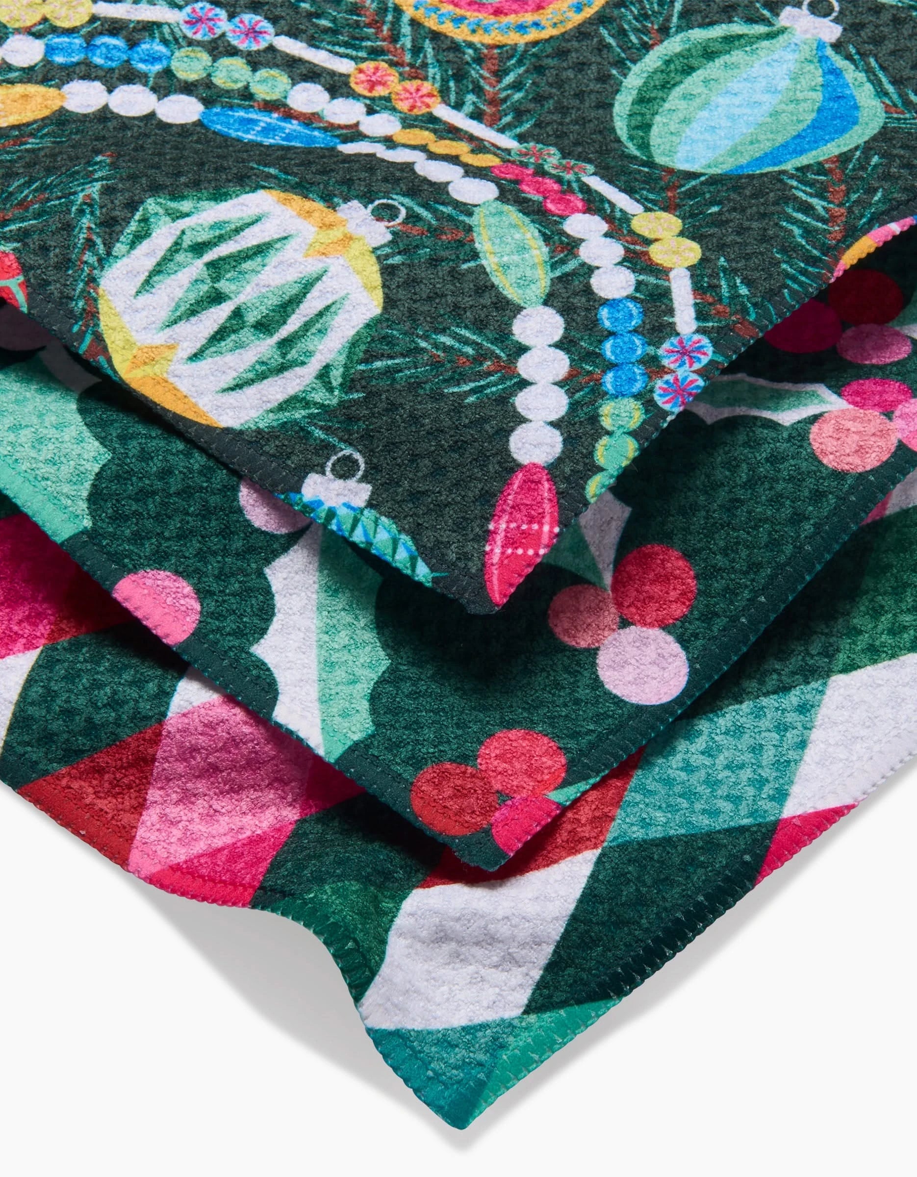 Three Retro Holly kitchen towels by Rebecca Bobko overlap: the top shows ornaments and beads, the middle has green, white, and red geometric shapes, and the bottom displays red holly berries—all in soft Geoweave fabric.