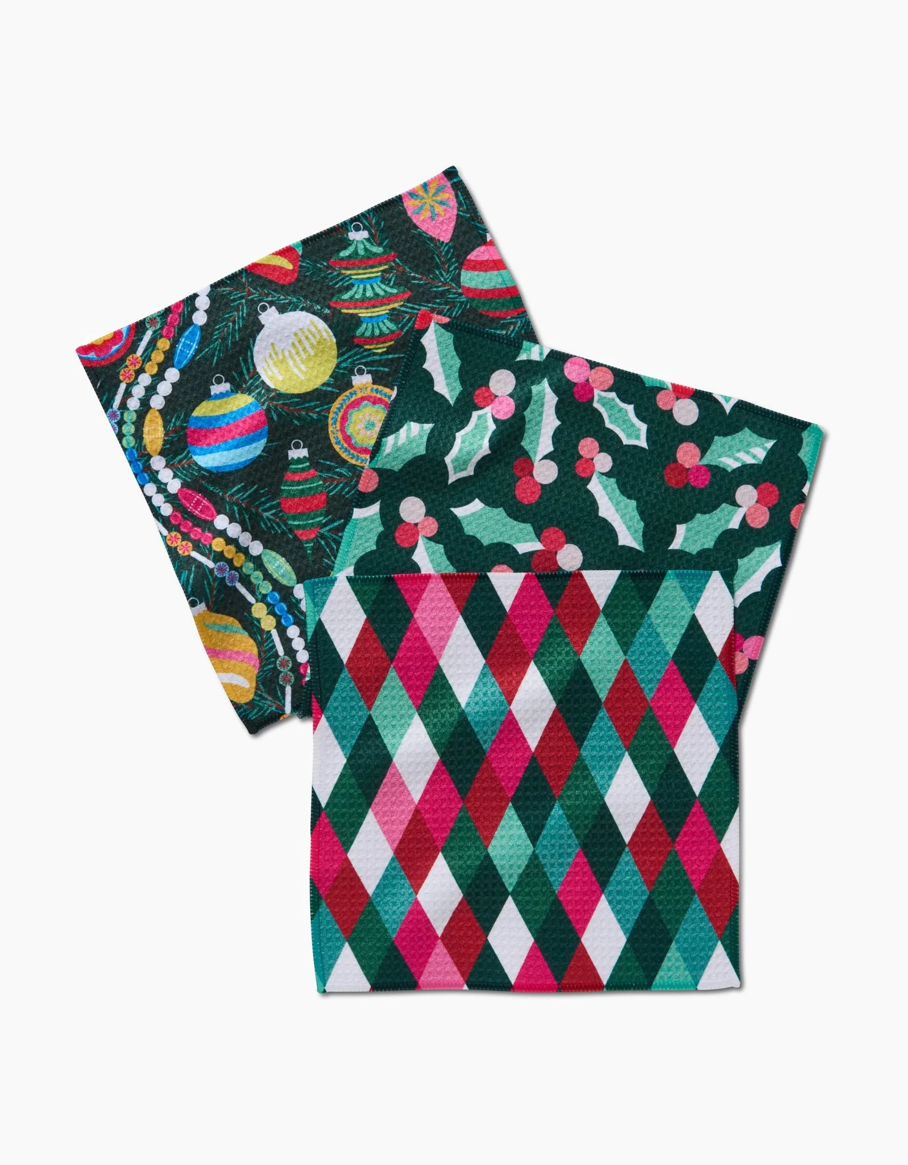 Two holiday-themed Rebecca Bobko Retro Holly Geoweave cloth napkins: one with ornaments and garland, another with holly leaves and berries, both tucked into a third napkin featuring a red, green, and white diamond pattern.