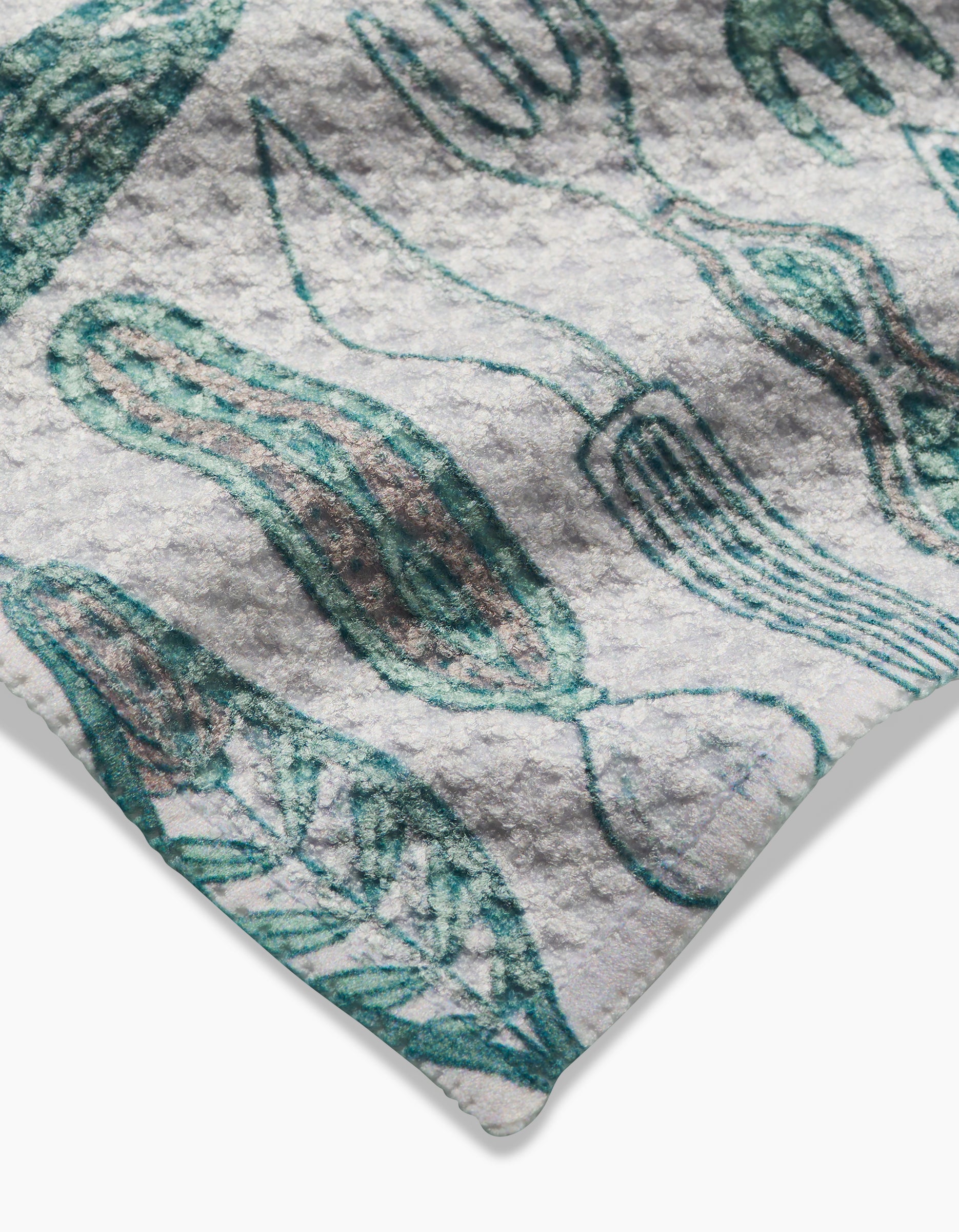 A close-up of the Romy Wares Geoweave dishcloth by Amy MacCready, showcasing a textured white surface with abstract green and brown plant and leaf illustrations, curved edges, and visible stitched seams.