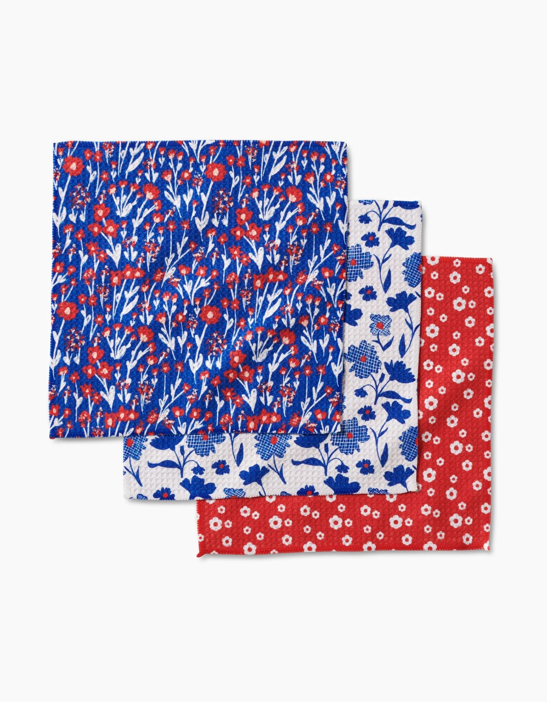 Three overlapping Royal Garden cloth napkins from Danelys Sidron are shown: the top features a blue background with red and white flowers, the middle is white with blue flowers, and the bottom is red with small white flowers. Made from recycled materials.