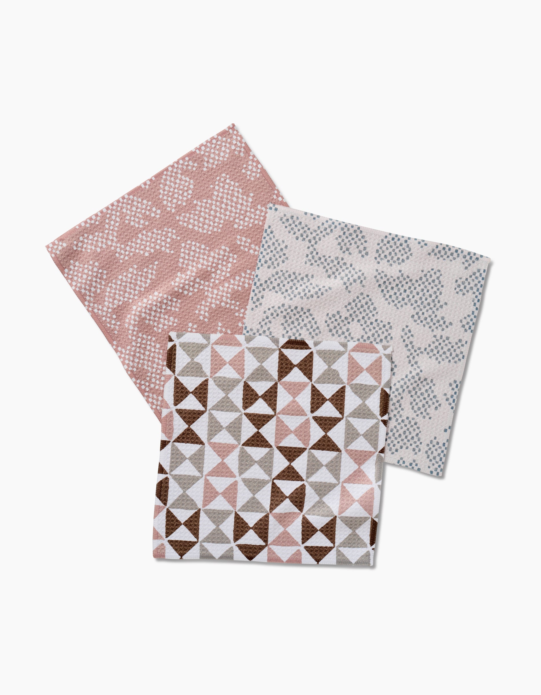 Studio Jeannot’s Rubix Pixie features three recycled cloth napkins: pink and white abstract, blue and white, and brown-pink-white geometric triangle designs, all overlapping on a white background.