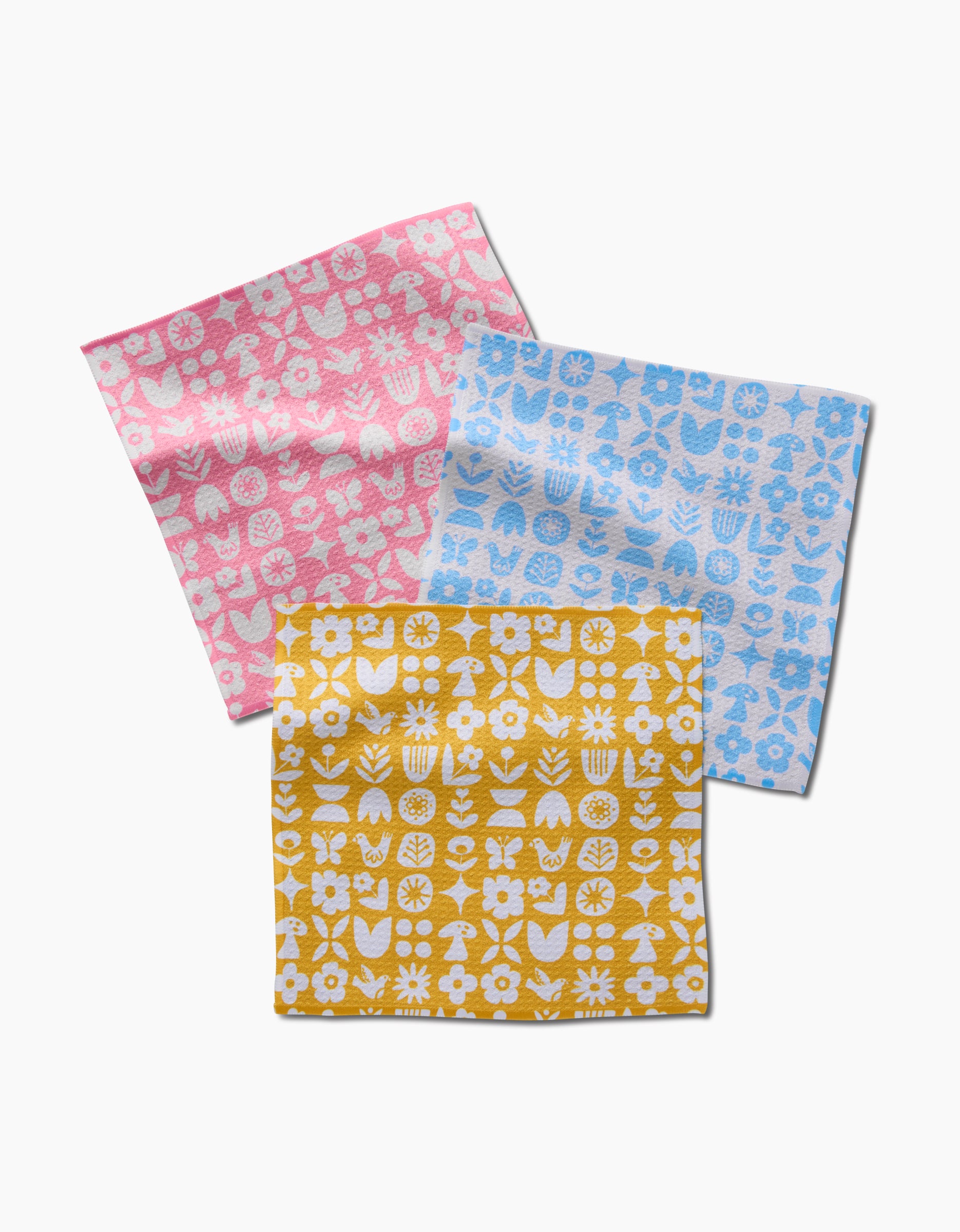 Three square Scandi Spring napkins by Kayla Ann in pink, blue, and yellow, each with white floral and plant patterns, are laid out overlapping on a white background to highlight their vibrant dishcloth designs.