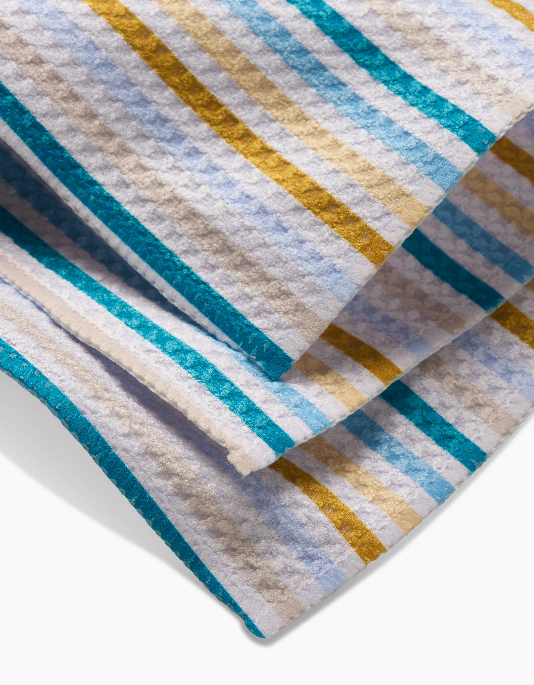 A close-up of the GEOMETRY Seaside Sunshine dishcloth, featuring textured Geoweave fabric with teal, golden yellow, white, and light blue stripes. The soft, slightly crinkled cloth is made from recycled materials.