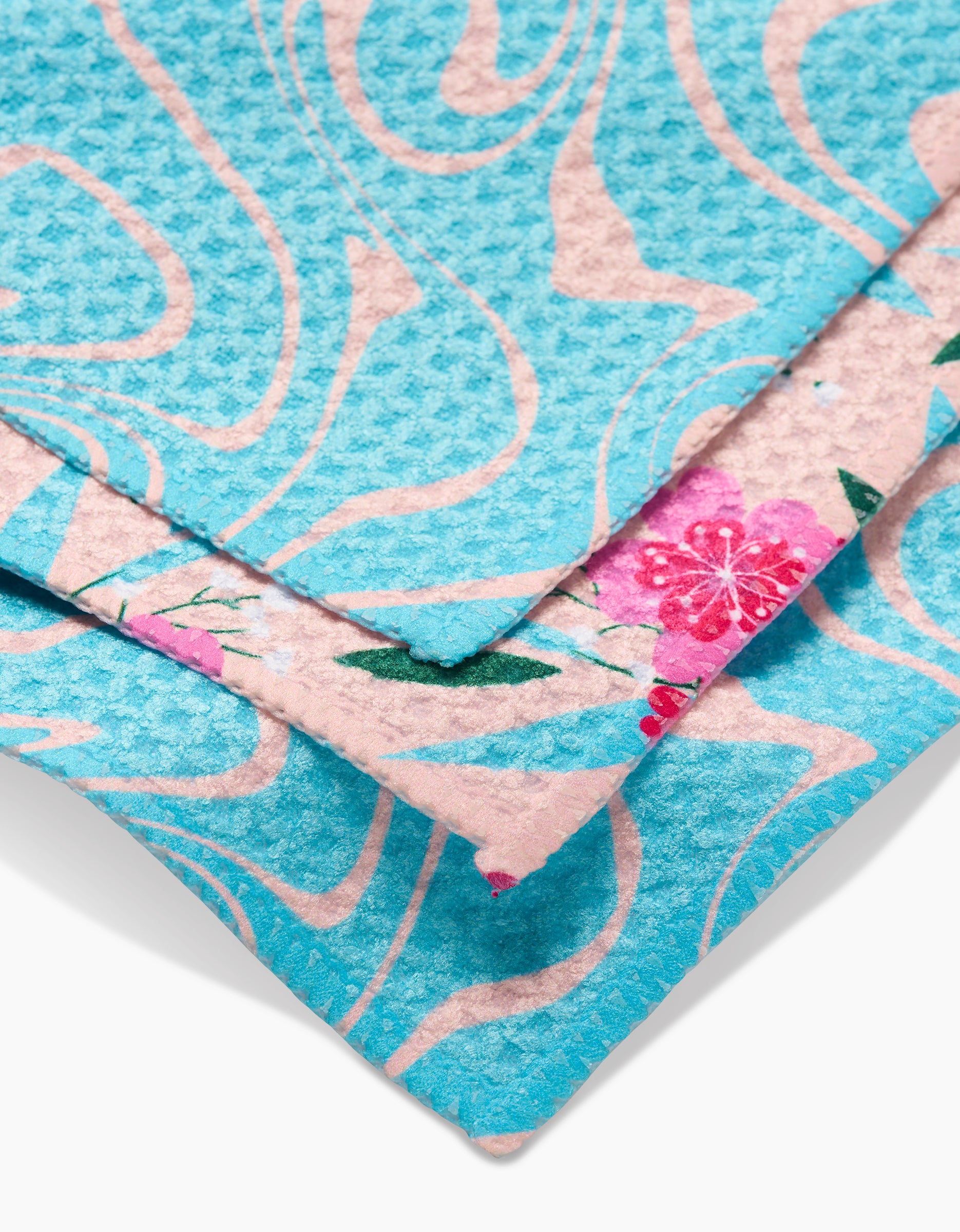 Two overlapping GEOMETRY dishcloths from the Sending Love collection: the top features a super-absorbent Geoweave blue swirl, while the bottom displays a pink floral pattern with green leaves.